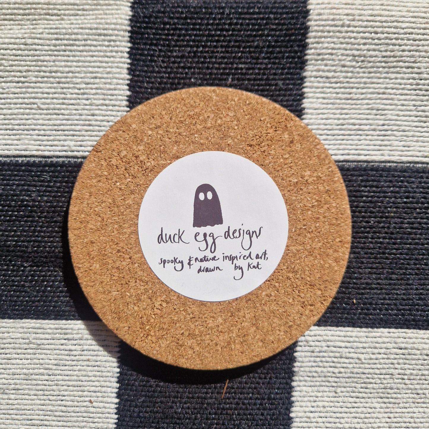 The Baking Ghost coaster by Duck Egg Designs features a simple ghost illustration and text “spooky & sassy inspired art, drawn by Kat” on a round cork base. Printed in the UK and shown on black-and-white checkered fabric.