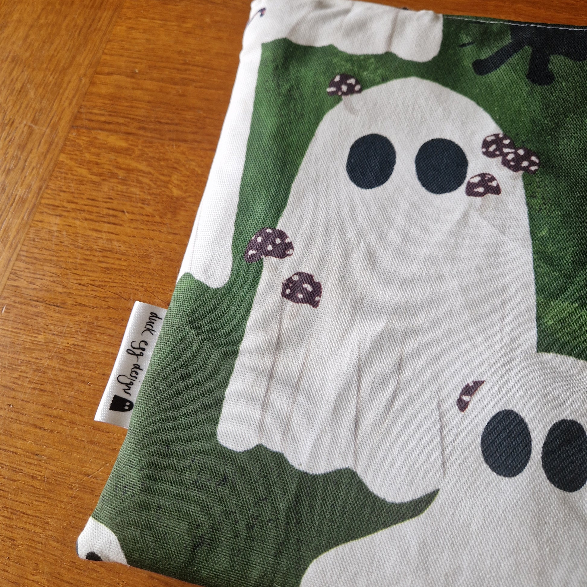 The A Haunting Of Ghosts Book Sleeve by Duck Egg Designs is a handmade pouch from recycled fabric, with white ghosts and tiny mushrooms, resting on wood. A small label reads “made by Emily.” Perfect for protecting your books!.