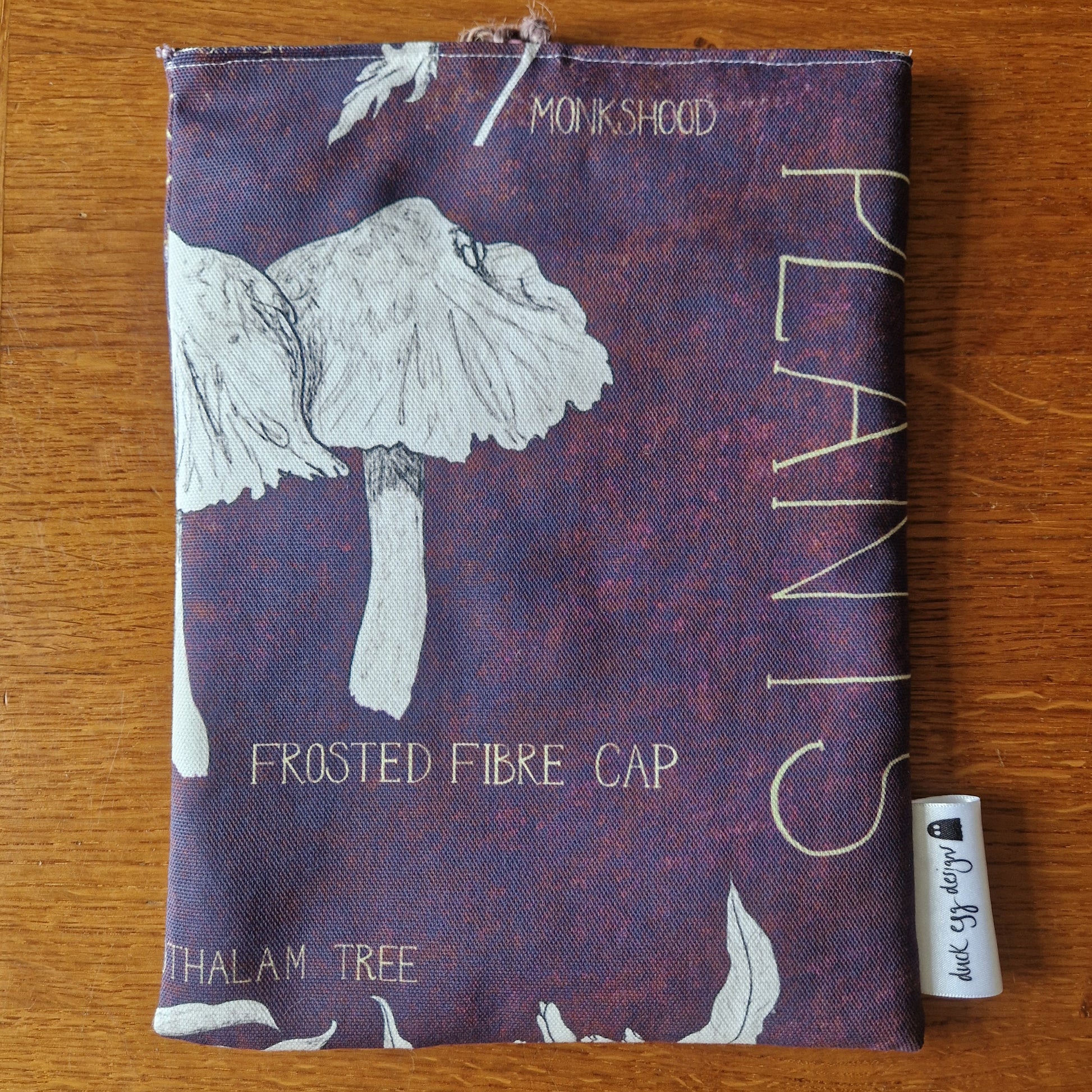 The Poison Plants Book Sleeve by Duck Egg Designs is a handmade pouch from upcycled fabric, featuring illustrated mushrooms and plants with labeled names on a maroon background, plus a small Made by Ivy&Em tag.