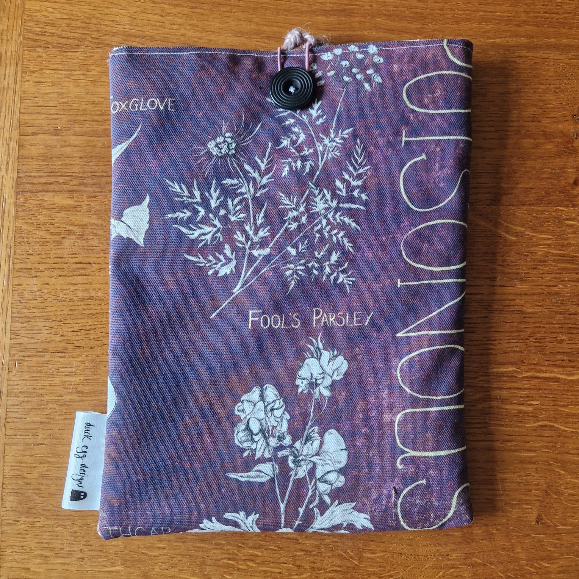 The Poison Plants Book Sleeve by Duck Egg Designs is a handmade pouch crafted from upcycled fabric, featuring botanical illustrations and plant labels on a purple background, finished with a black button and tag.