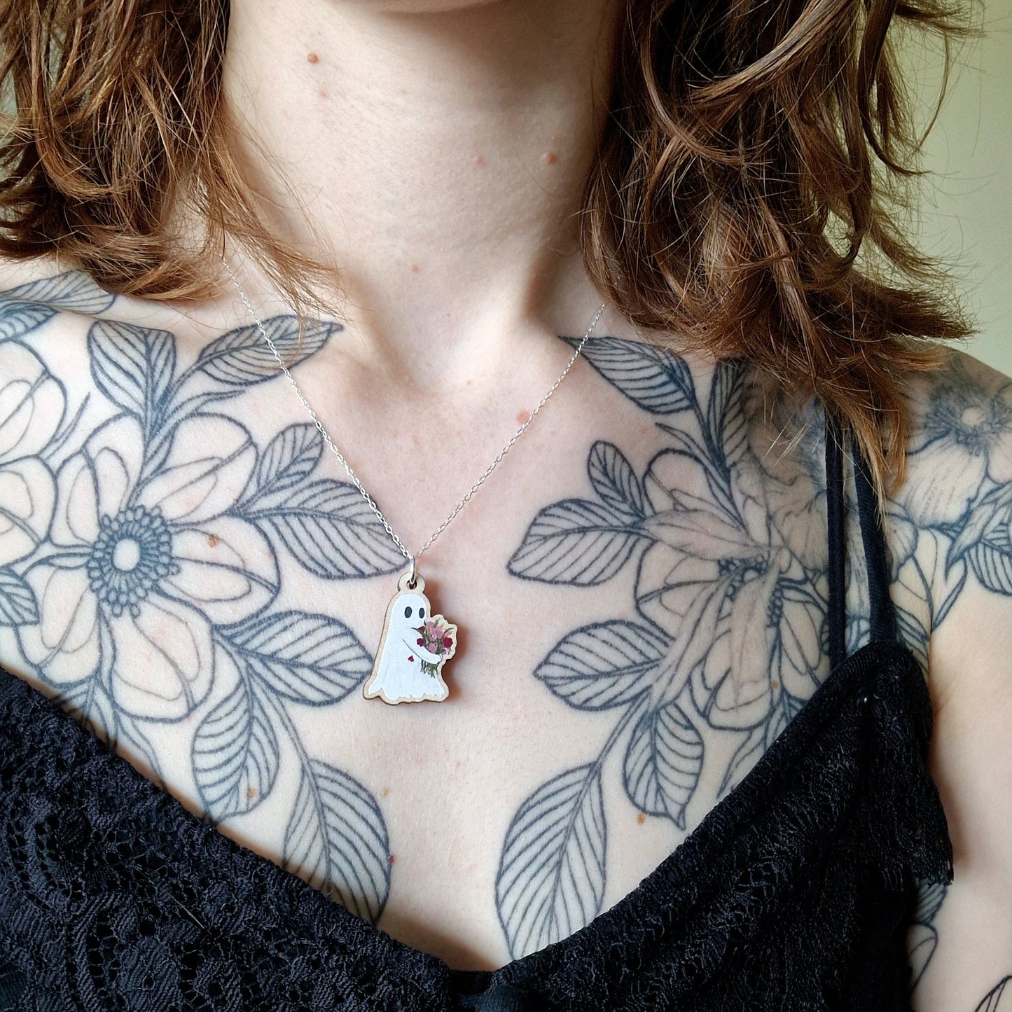 Person wearing a black top with floral tattoos on shoulders and chest, necklace with a pendant featuring a ghost holding a bunch of flowers.
