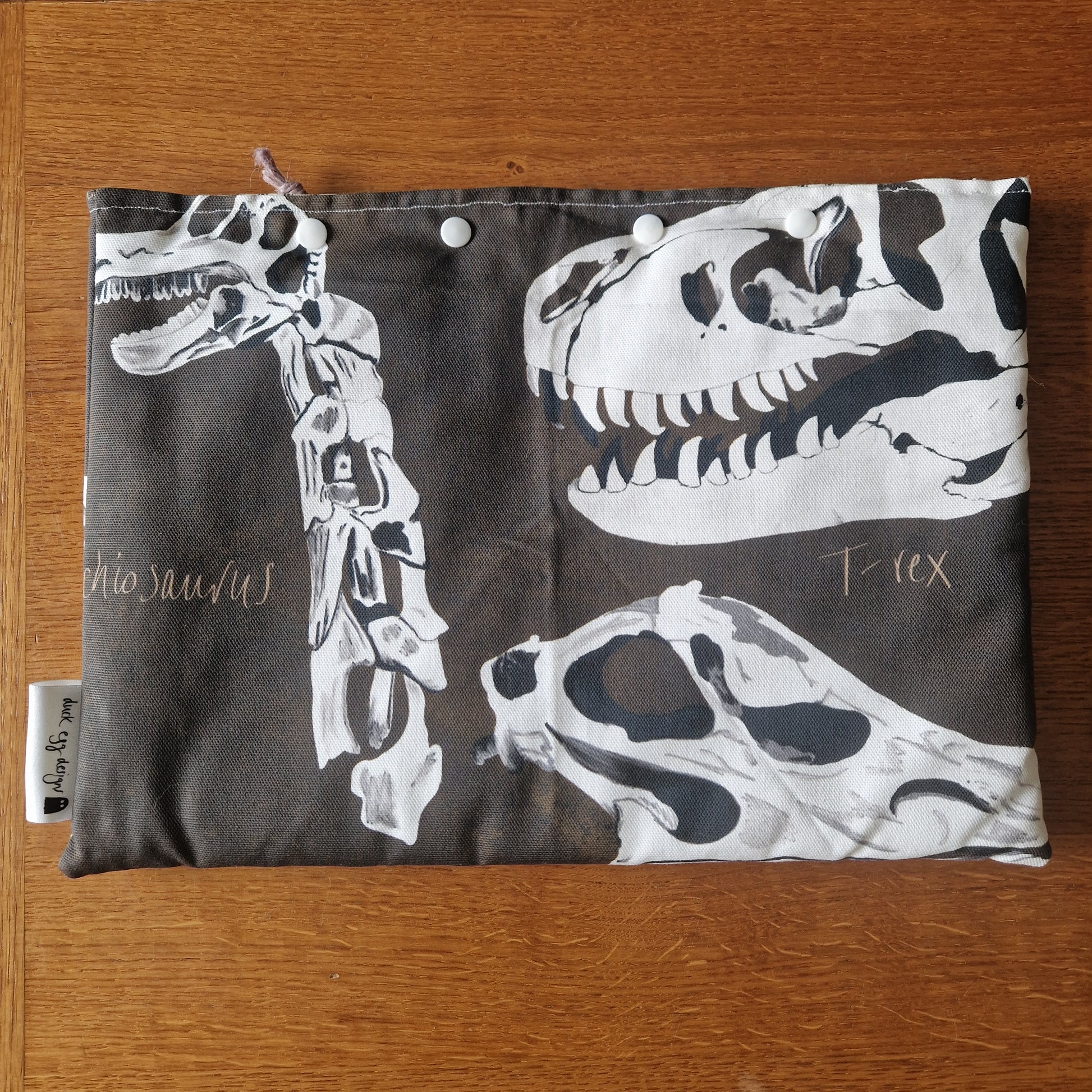 The Dinosaur Bones Small Laptop/Tablet Sleeve by Duck Egg Designs is a handmade fabric pouch with white snap buttons, featuring white dinosaur skeletons—including a T. rex skull—and cursive “dinosaurs” and “T rex” on a dark background.