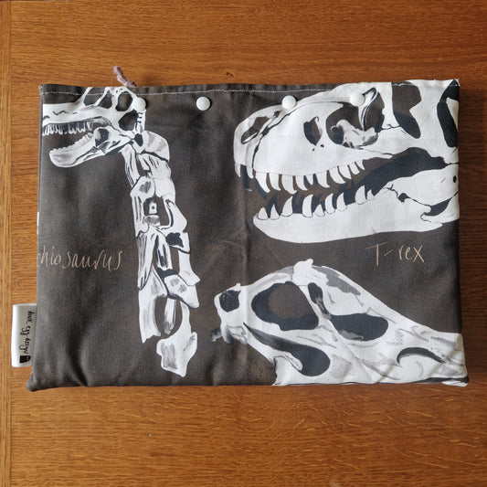 The Dinosaur Bones Small Laptop/Tablet Sleeve by Duck Egg Designs is a handmade fabric pouch with white snap buttons, featuring white dinosaur skeletons—including a T. rex skull—and cursive “dinosaurs” and “T rex” on a dark background.