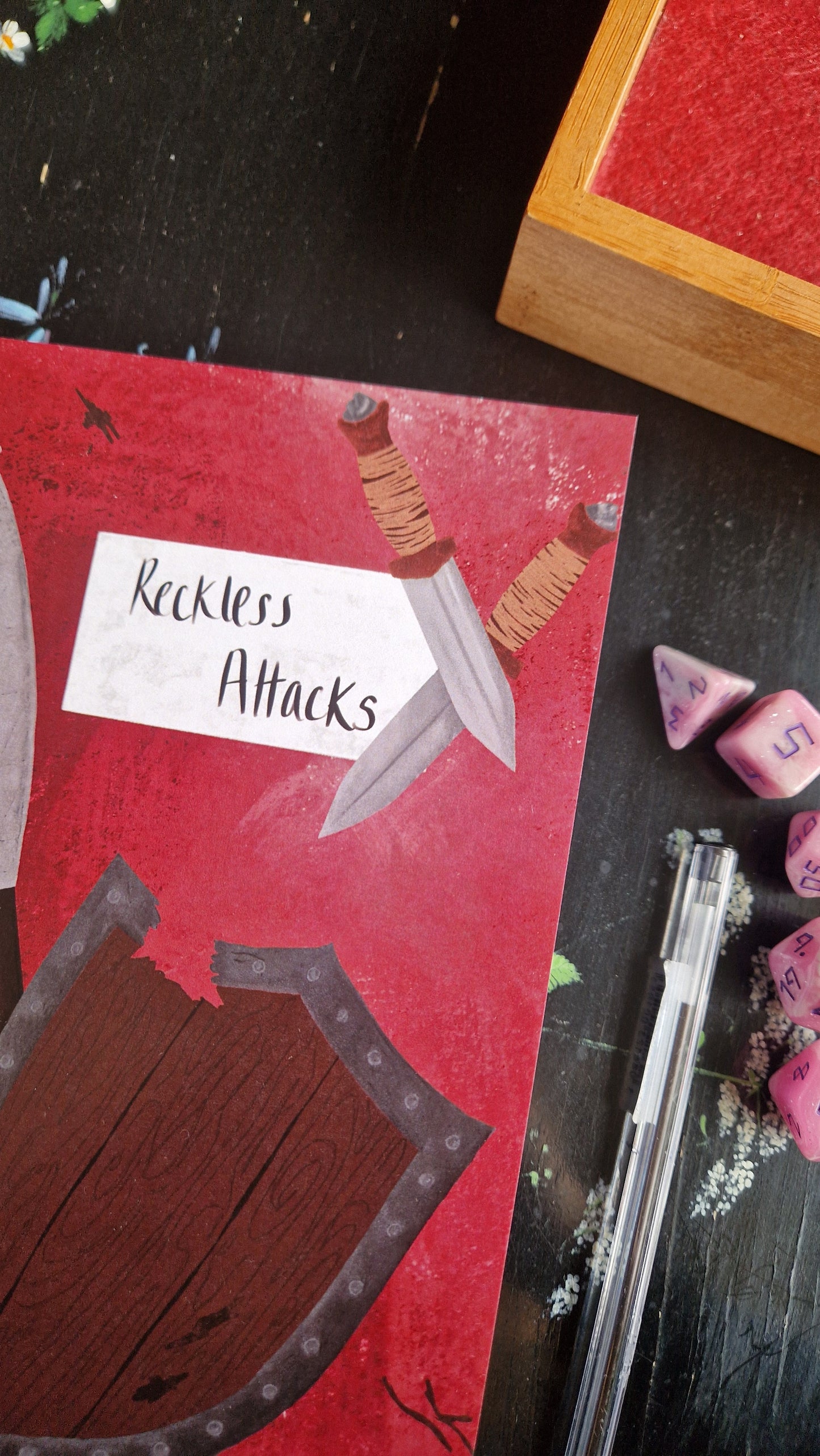 The Duck Egg Designs Reckless Attack Notebook features red cover art with two daggers, a wooden shield, and handwritten text. Shown on a dark tabletop with a pen, pink polyhedral dice, campaign notes, and a wooden box.