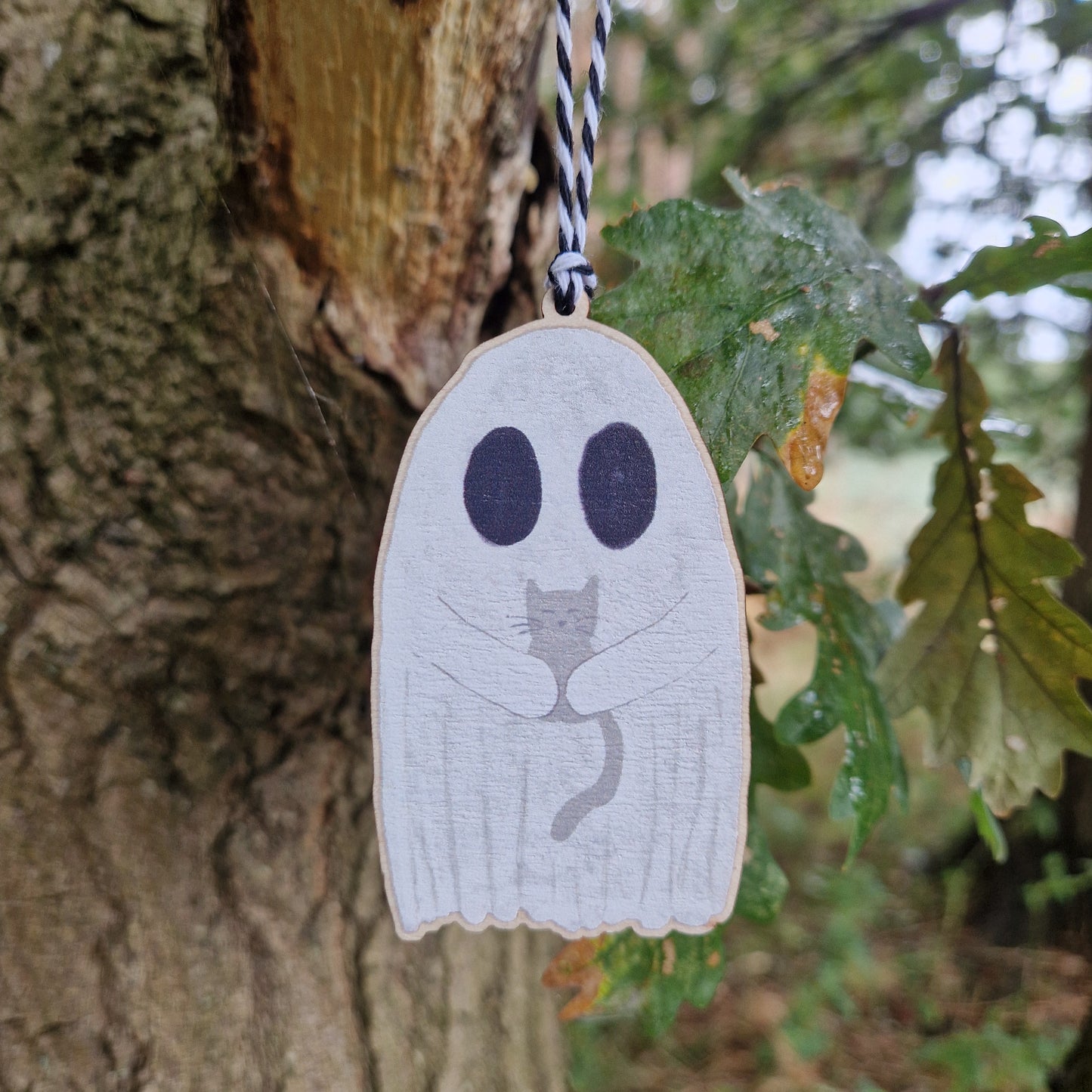 The Ghost With Cat Hanging Wooden Decoration by Duck Egg Designs features a ghost holding a gray cat, suspended by a black-and-white string. Green oak leaves and textured bark provide a charming backdrop for this unique ornament.