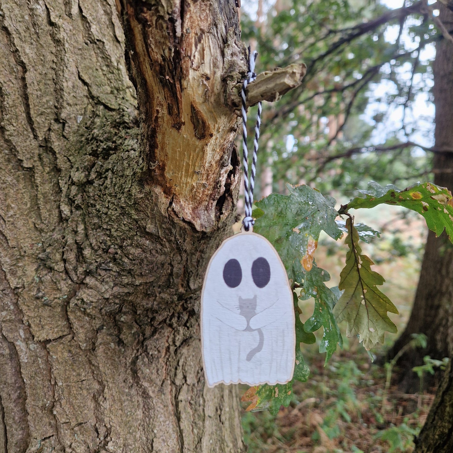 The Ghost With Cat Hanging Wooden Decoration by Duck Egg Designs features a black cat illustration and is displayed as a wooden charm hanging from a tree trunk in the woods.