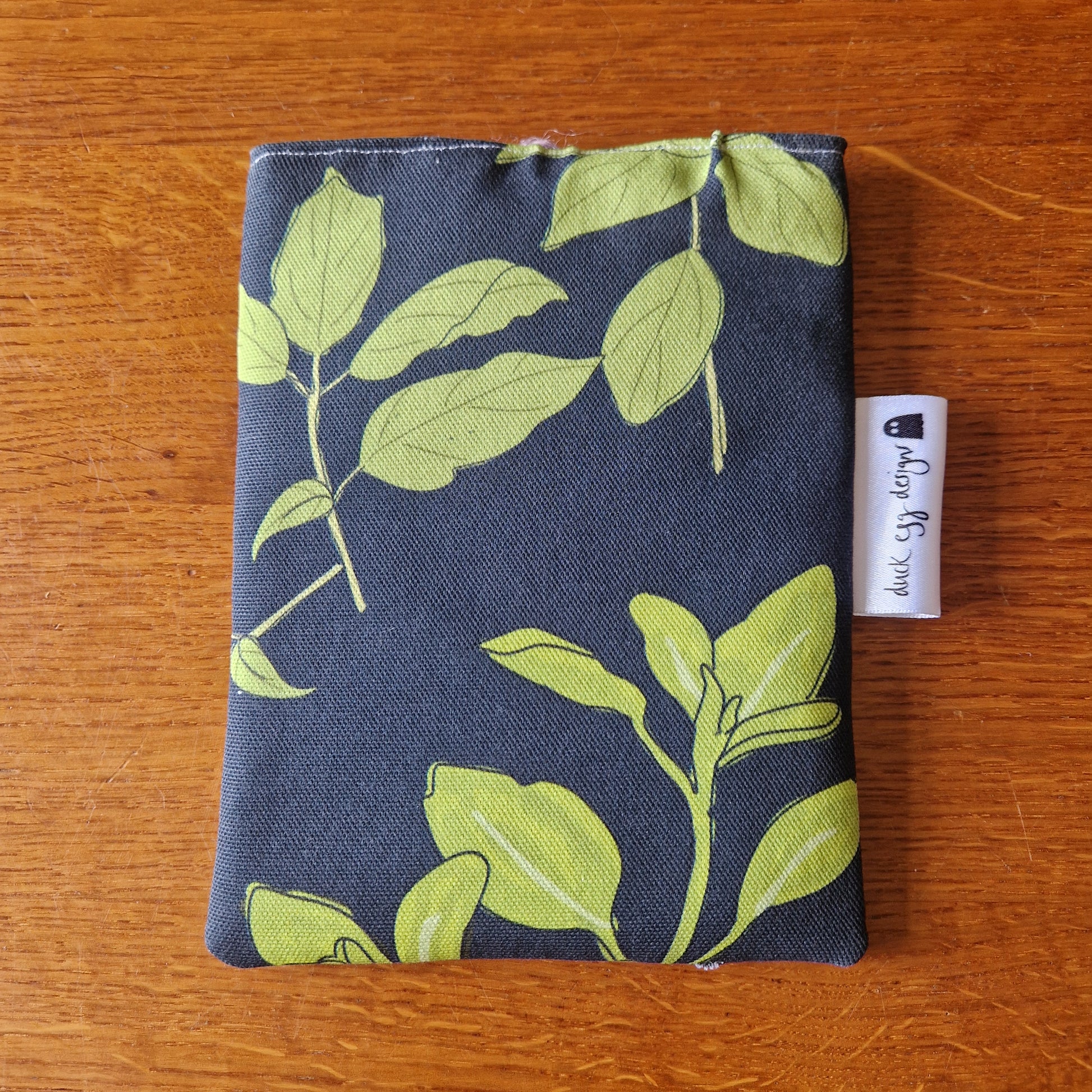 The Common Herbs Kindle Pouch by Duck Egg Designs features a green leaf pattern on a dark background. A white tag with black text is attached to the handmade sleeve, which rests on a wooden surface.