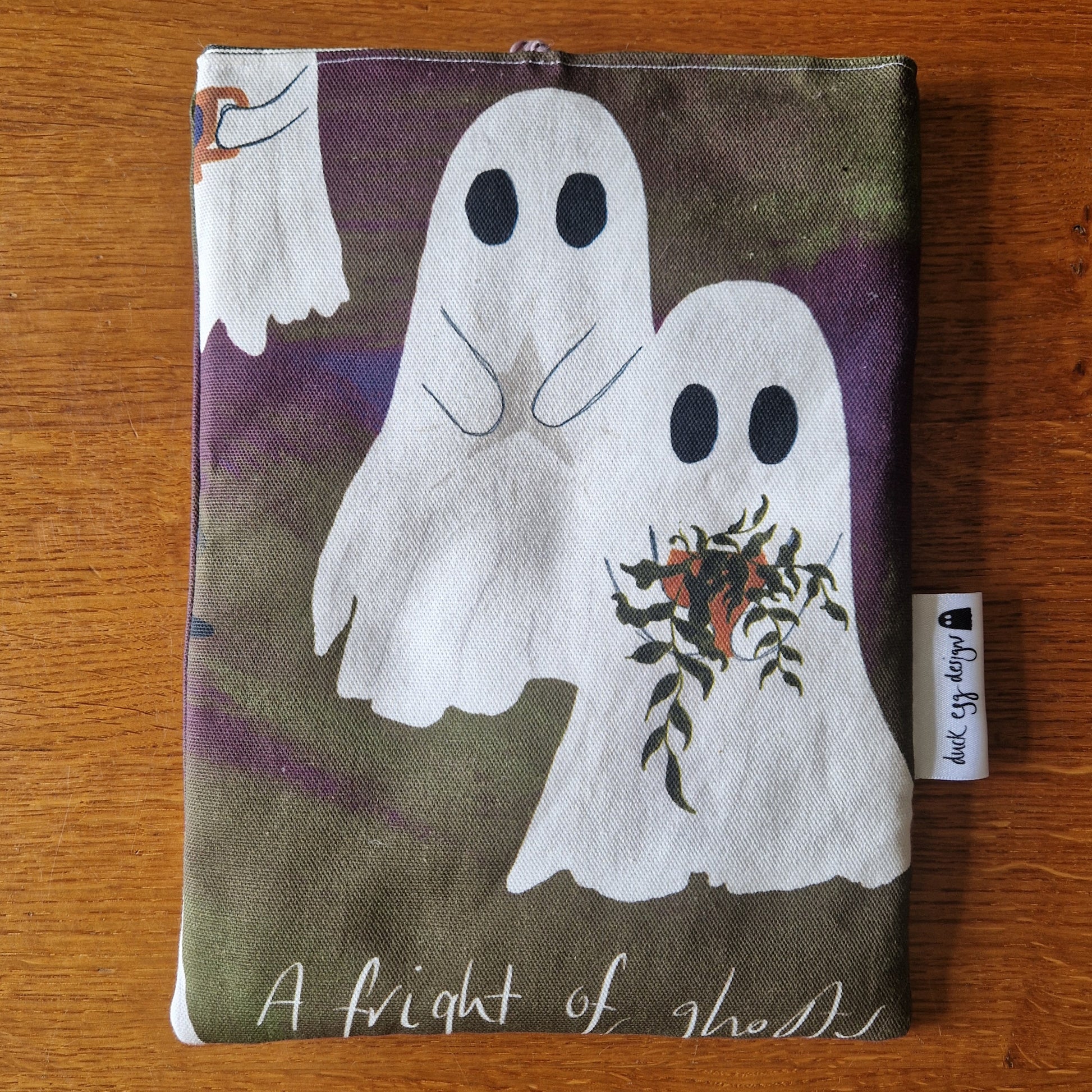The A Fright Of Ghosts Book Sleeve by Duck Egg Designs is a reusable pouch made from upcycled fabric, featuring two cartoon ghosts—one with dried flowers—on a dark pattern with A fright of ghosts at the bottom.