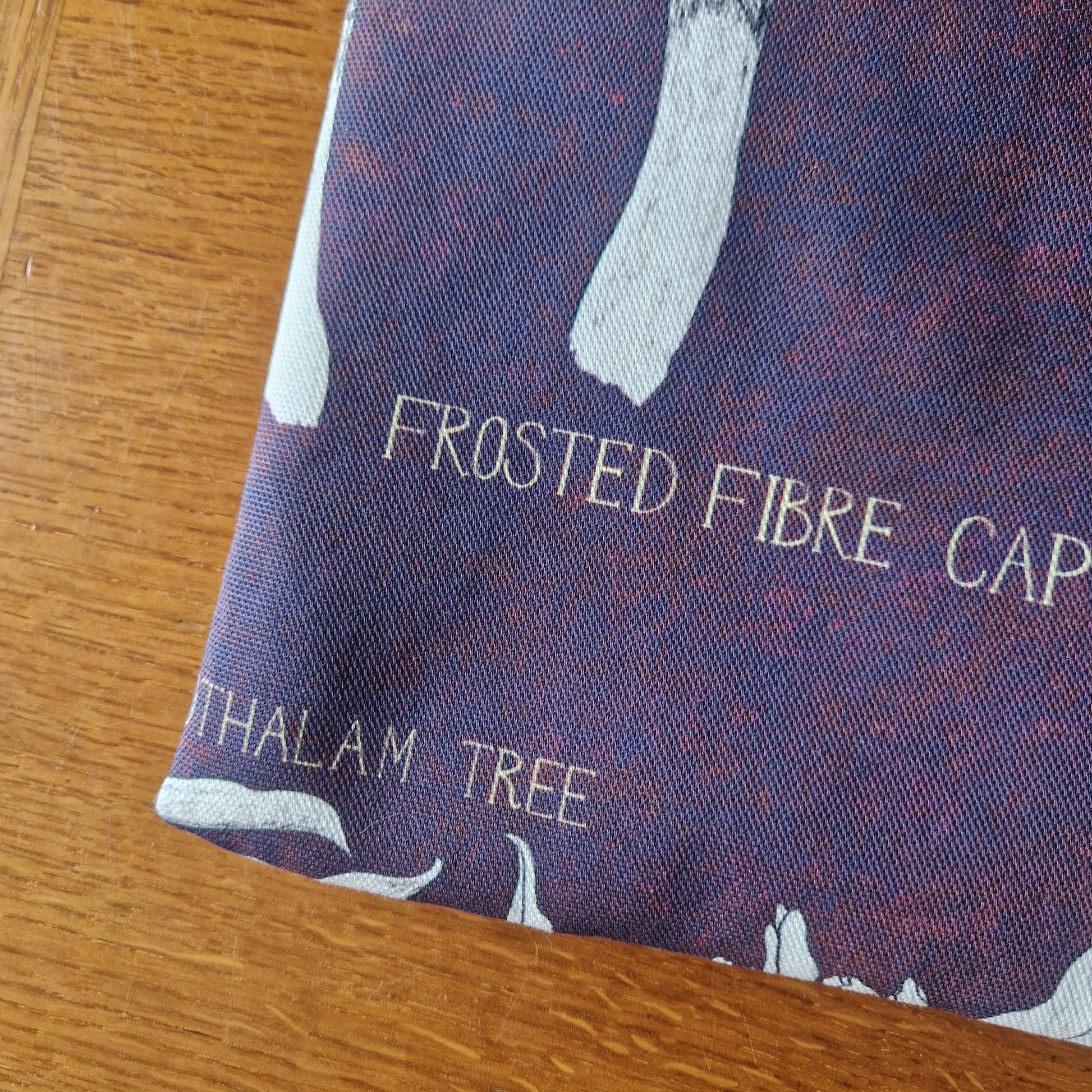 A close-up of Duck Egg Designs Poison Plants Book Sleeve shows dark blue upcycled fabric with white botanical illustrations and text, including FROSTED FIBRE and THALAM TREE, laid on a wooden surface.