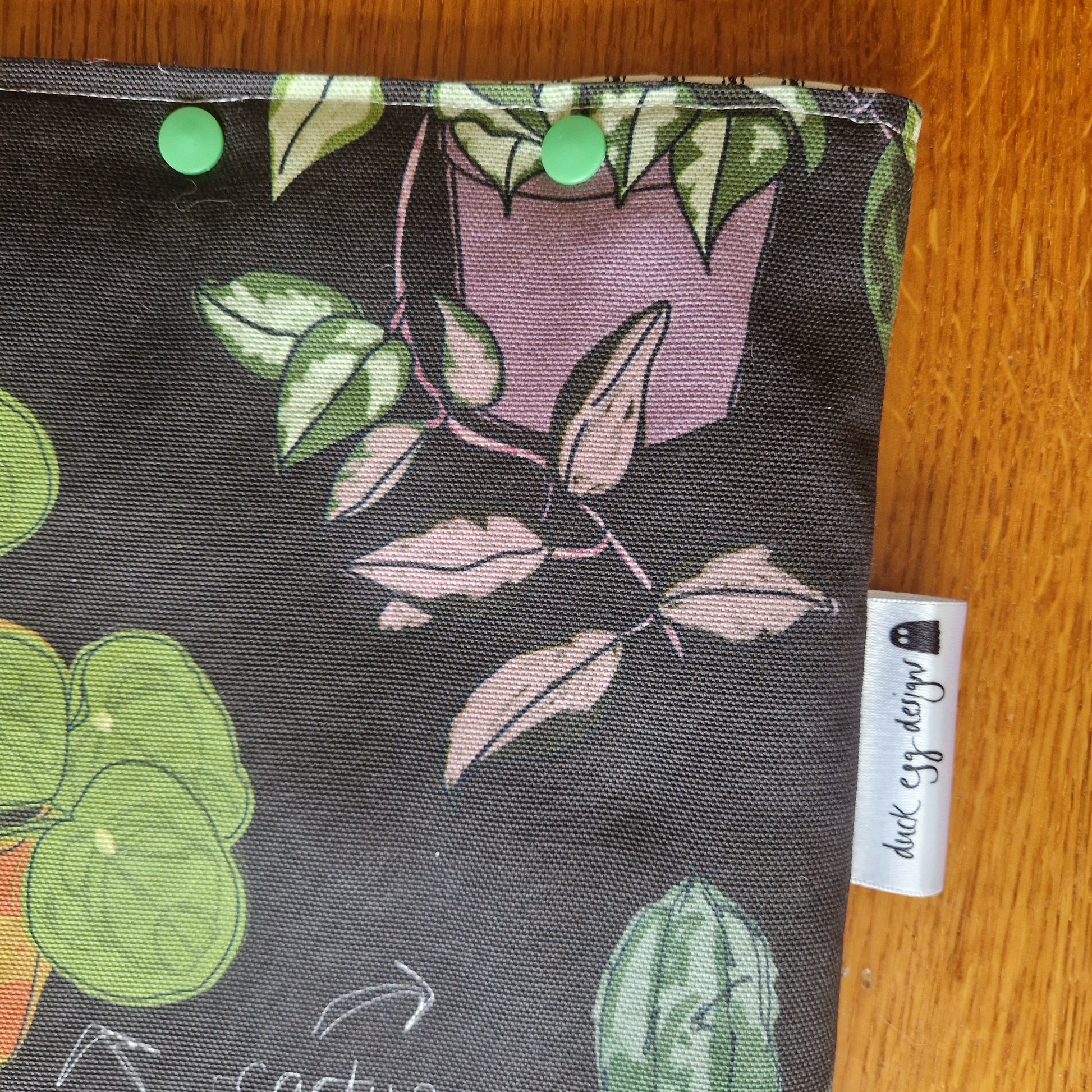 A close-up of Duck Egg Designs’ Common Houseplants Small Laptop/Tablet Sleeve, handmade from recycled fabric with a colorful plant print, green snap buttons, and a side label, displayed on a wooden surface.