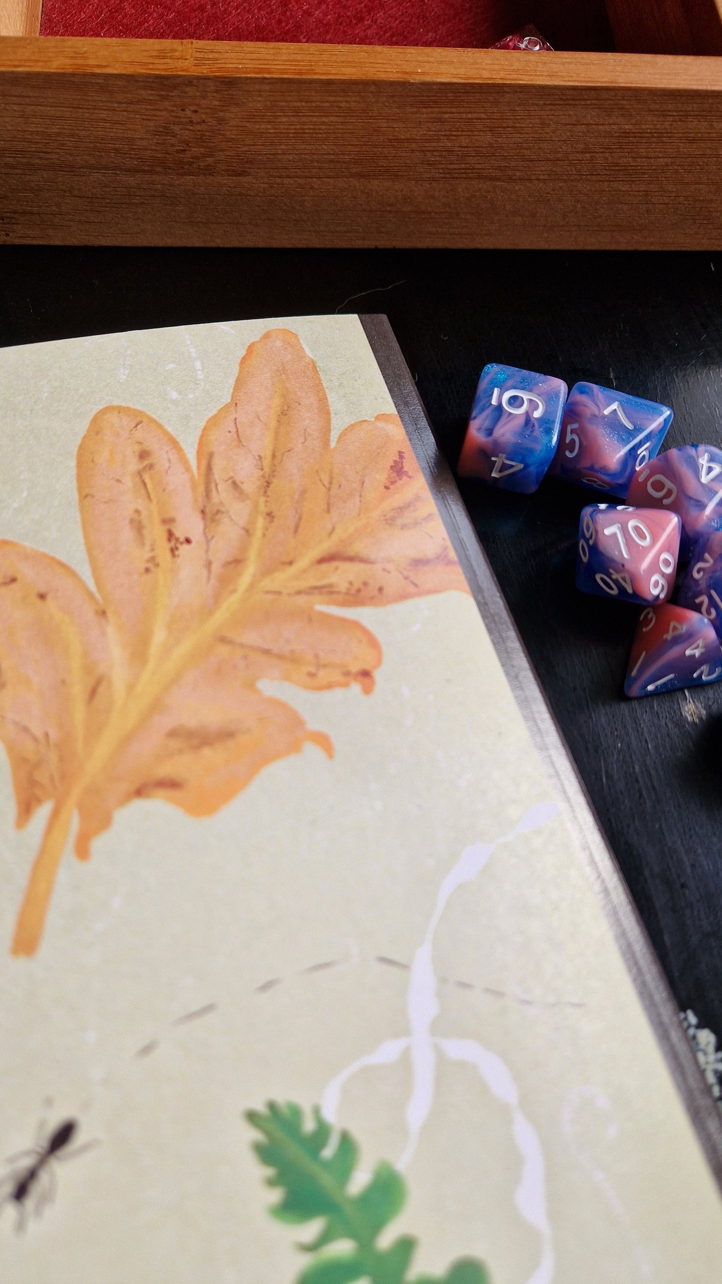 A close-up of the Duck Egg Designs Ways Of The Wood Notebook, featuring a brown leaf illustration, sits beside blue and pink polyhedral dice on a dark surface with a wooden tray in the background.