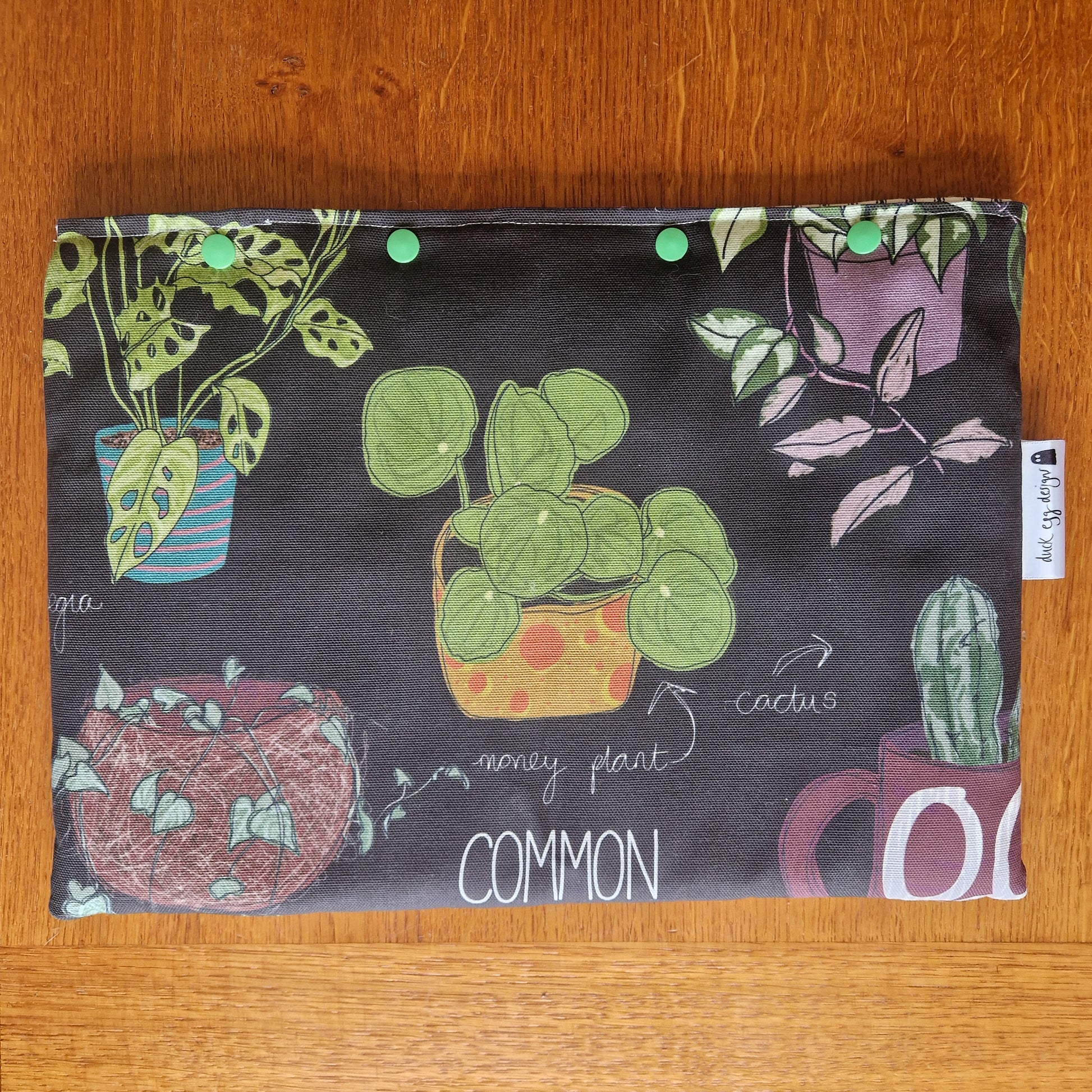 The Common Houseplants Small Laptop/Tablet Sleeve by Duck Egg Designs features colorful potted plant illustrations, including a money plant and cactus with handwritten labels, handmade from recycled fabric and finished with green snap buttons.
