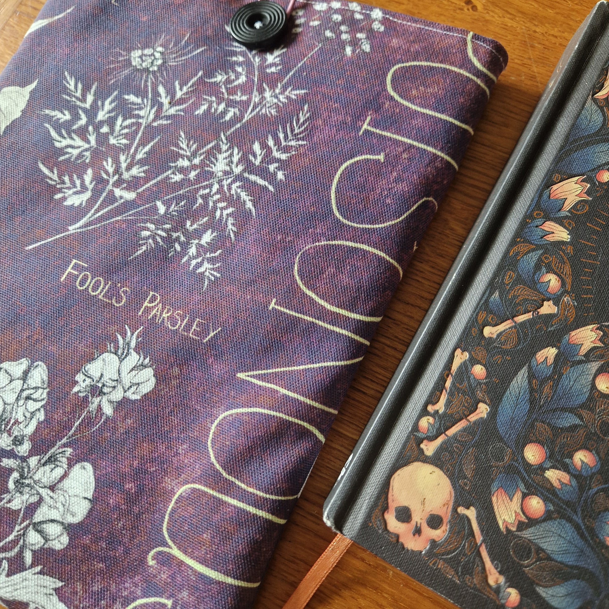 A close-up of two books in Duck Egg Designs Poison Plants Book Sleeve—one sleeve with a Fools Parsley motif, the other showing skulls, bones, and leaves on upcycled fabric.