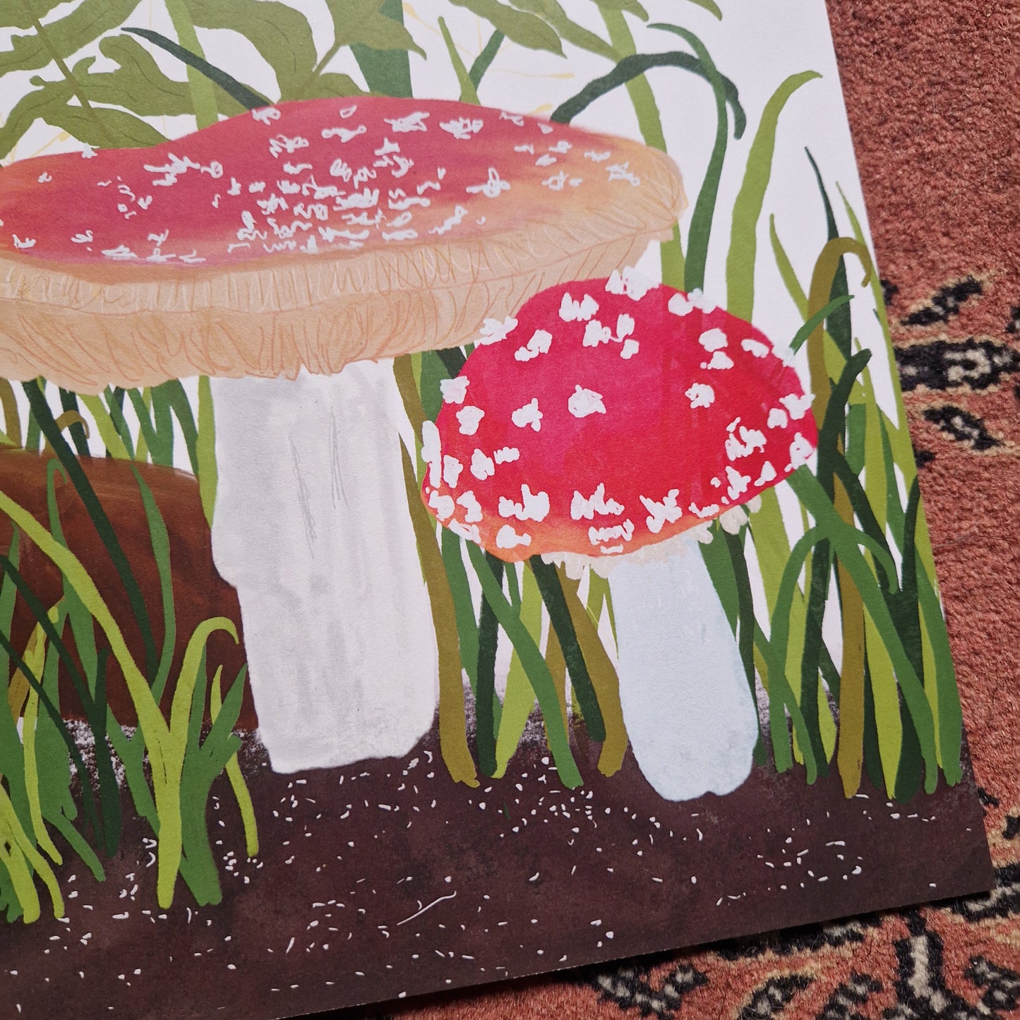 A close up of the bottom right of the print showcasing the detail in the artwork. You can see two of the fly agaric toadstools as well as green grasses and the brown soil. To the right and bottom of the print you can see a red textured patterned background.