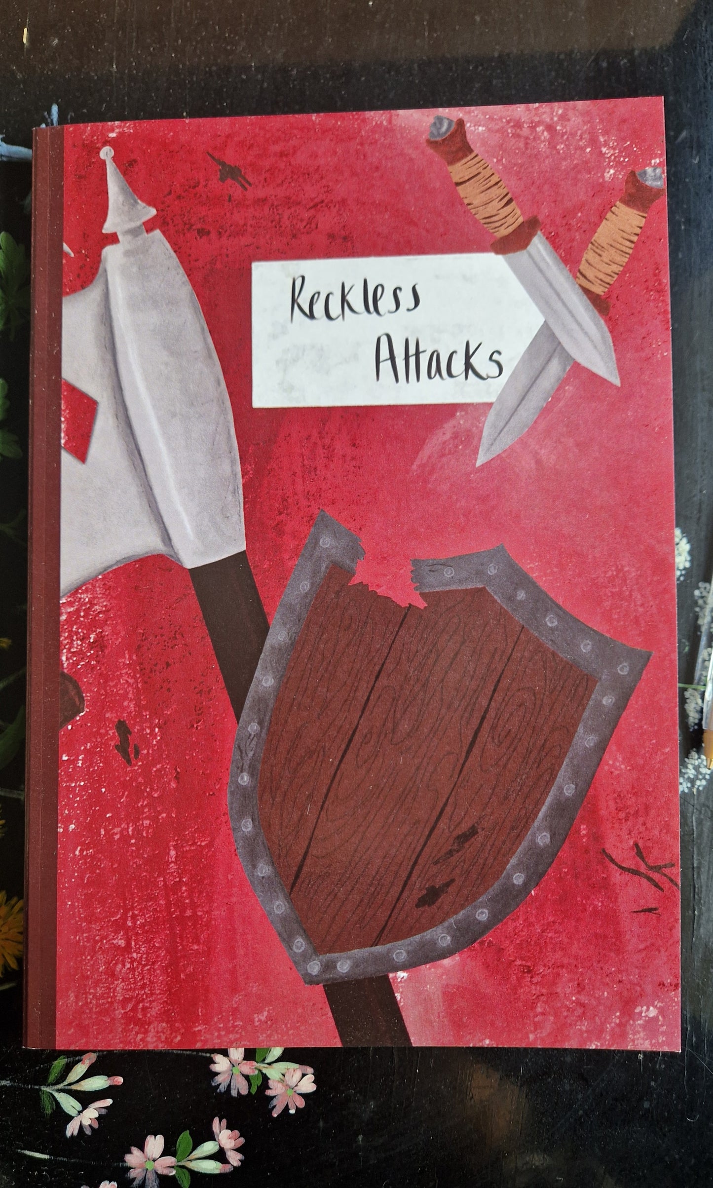 The Reckless Attack Notebook by Duck Egg Designs features a textured red cover with an axe, two knives, and a cracked wooden shield—ideal for barbarian notes or campaigns. Printed on recycled paper.