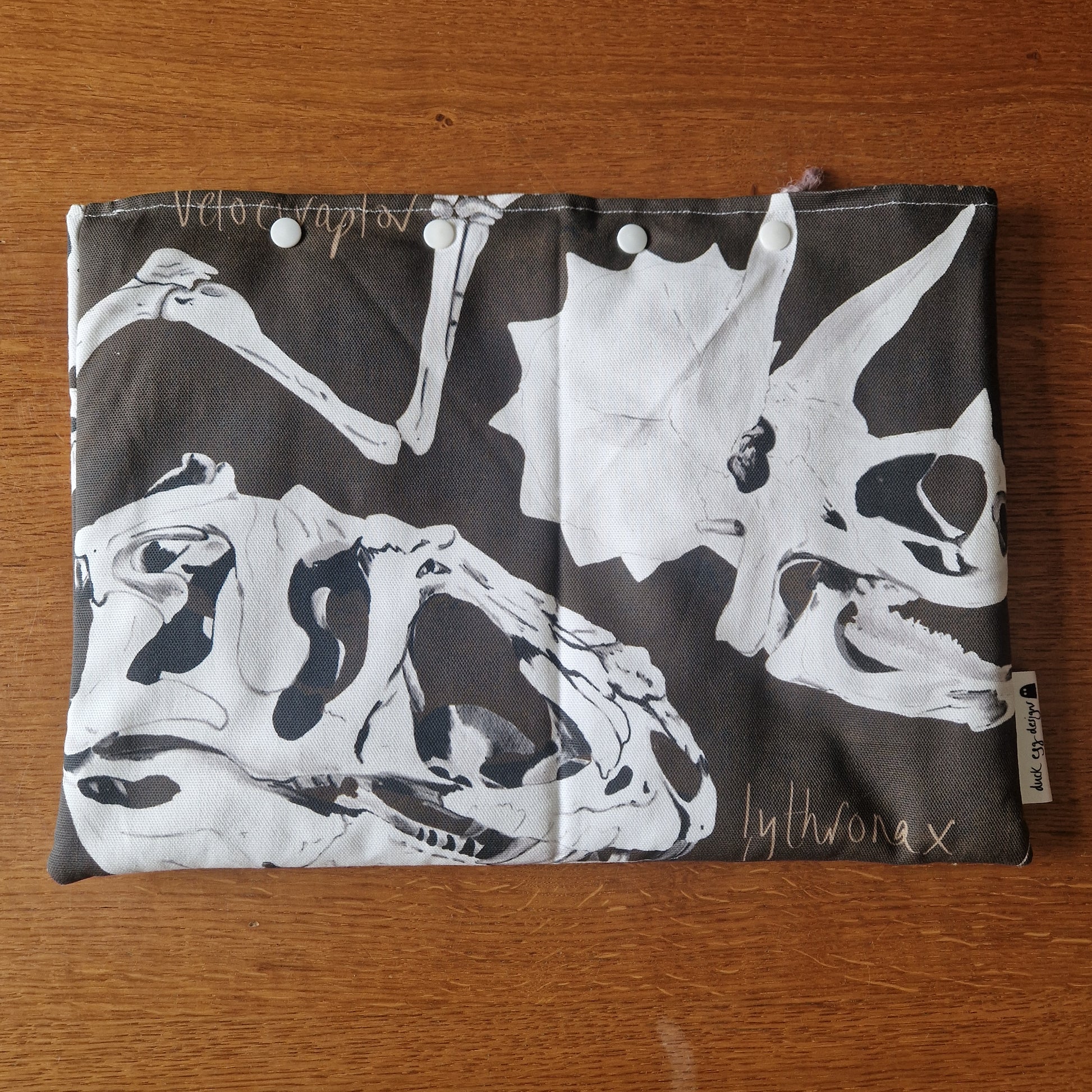 The Dinosaur Bones Small Laptop/Tablet Sleeve by Duck Egg Designs features a black and white dinosaur skull print, white snap buttons, and a side fabric label—handmade from recycled fabric.