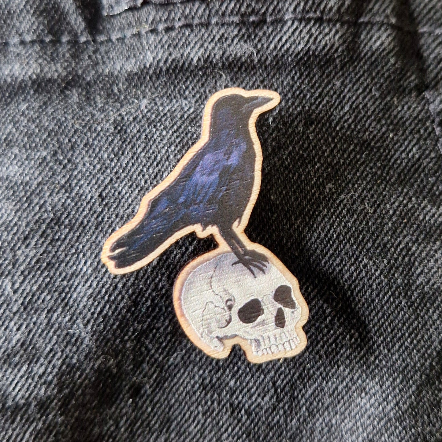 The Crow and Skull Pin Badge by Duck Egg Designs features a black raven perched on a human skull, all crafted in the UK and attached to textured black fabric.