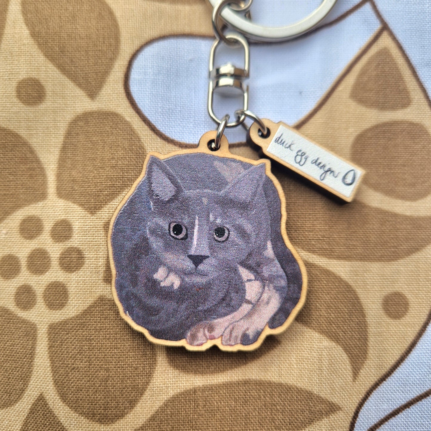 The Luna Grey Cat Keyring by Duck Egg Designs features an illustrated grey cat lying down, attached to a small sustainable wood tag. Presented on a brown and white floral-patterned fabric background. UK made.
