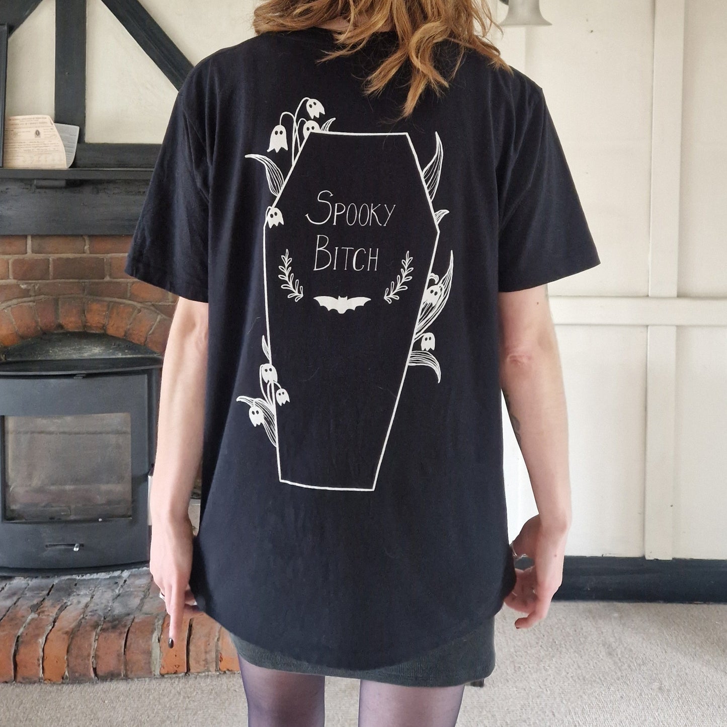 Person wearing a black t-shirt with white text and graphics, standing in a room with a door, a wall and a fireplace.