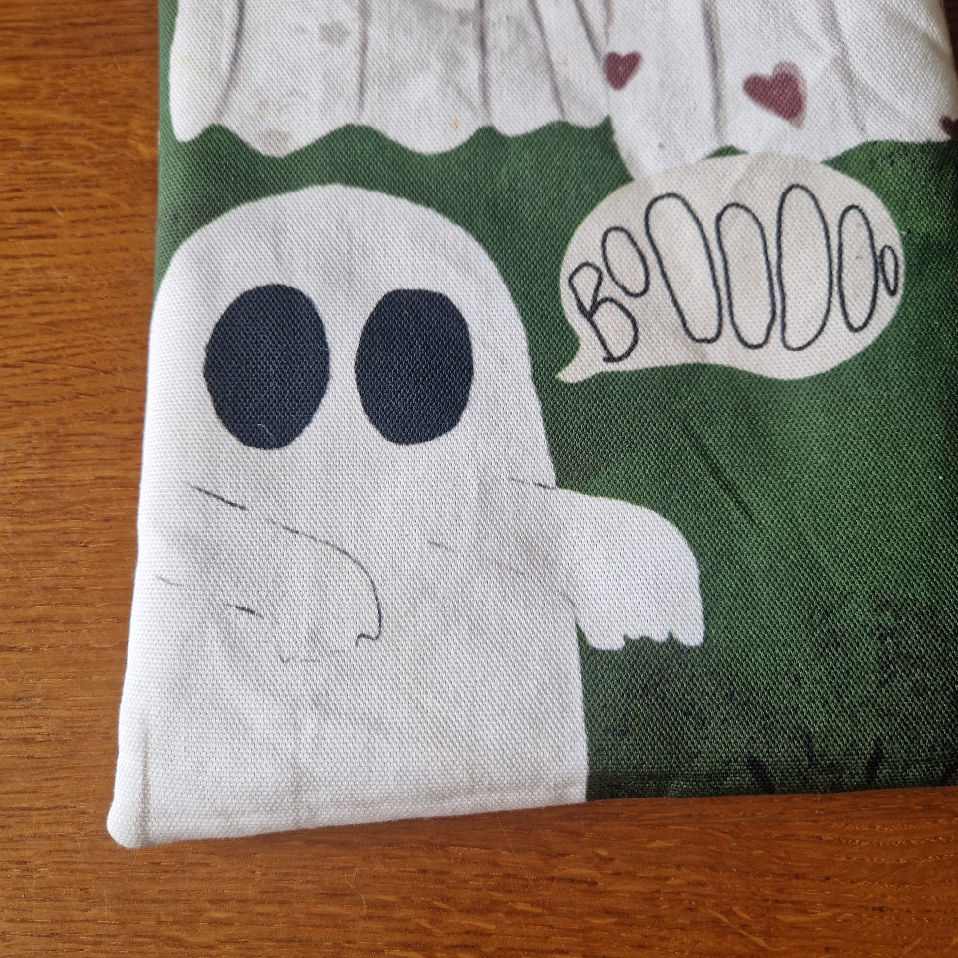 The A Haunting Of Ghosts Book Sleeve by Duck Egg Designs features a simple white cartoon ghost with big black eyes, red hearts, and a Booooo speech bubble, all printed on eco-friendly green recycled fabric—perfect for protecting your books.