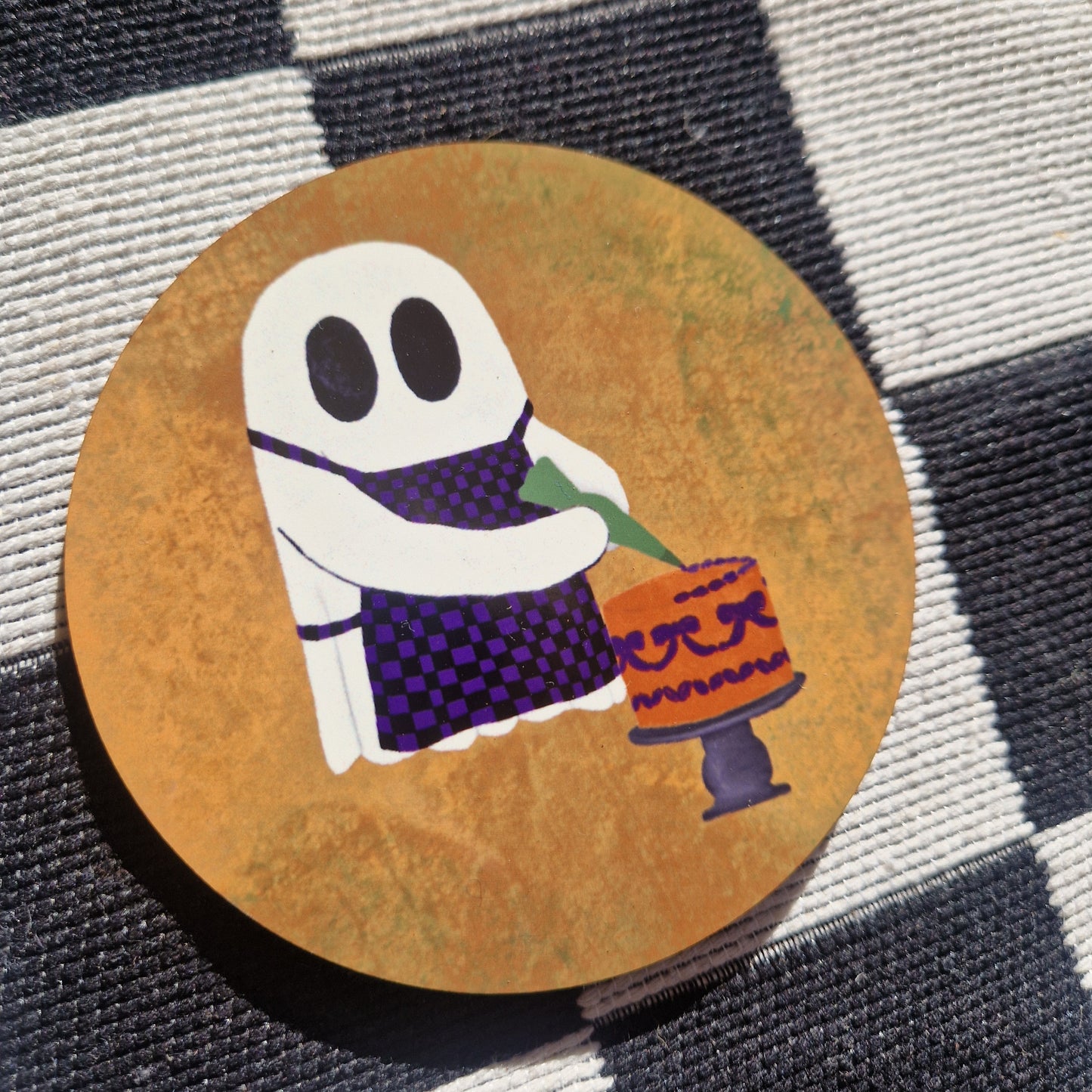 The Baking Ghost coaster by Duck Egg Designs features a cartoon ghost in a purple checkered apron decorating a cake with orange and purple icing, set against a gold background. Made in the UK with a cork base, it’s shown on a black and white checkered surface.