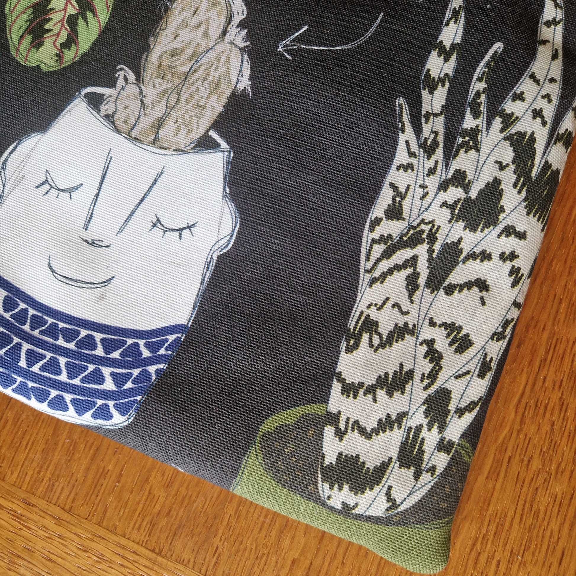 A close-up of Duck Egg Designs Common Houseplants Small Laptop/Tablet Sleeve, handmade from recycled fabric and featuring potted plant illustrations—a smiling cactus and a tall patterned succulent—resting on wood.