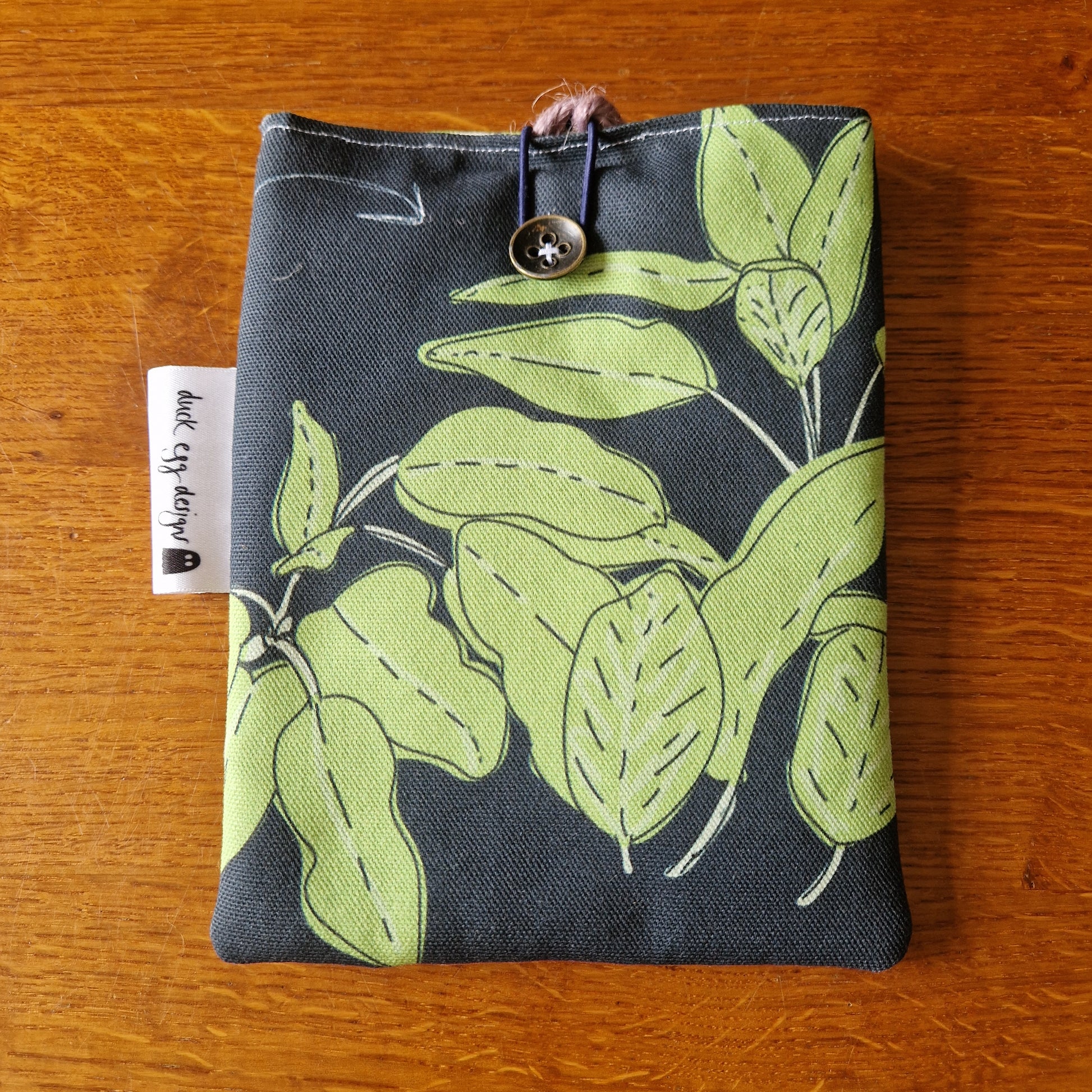 The Common Herbs Kindle Pouch by Duck Egg Designs is a handmade case with light green leaf patterns on a dark background, closed by a button and loop, and features a white tag with text on the left side.