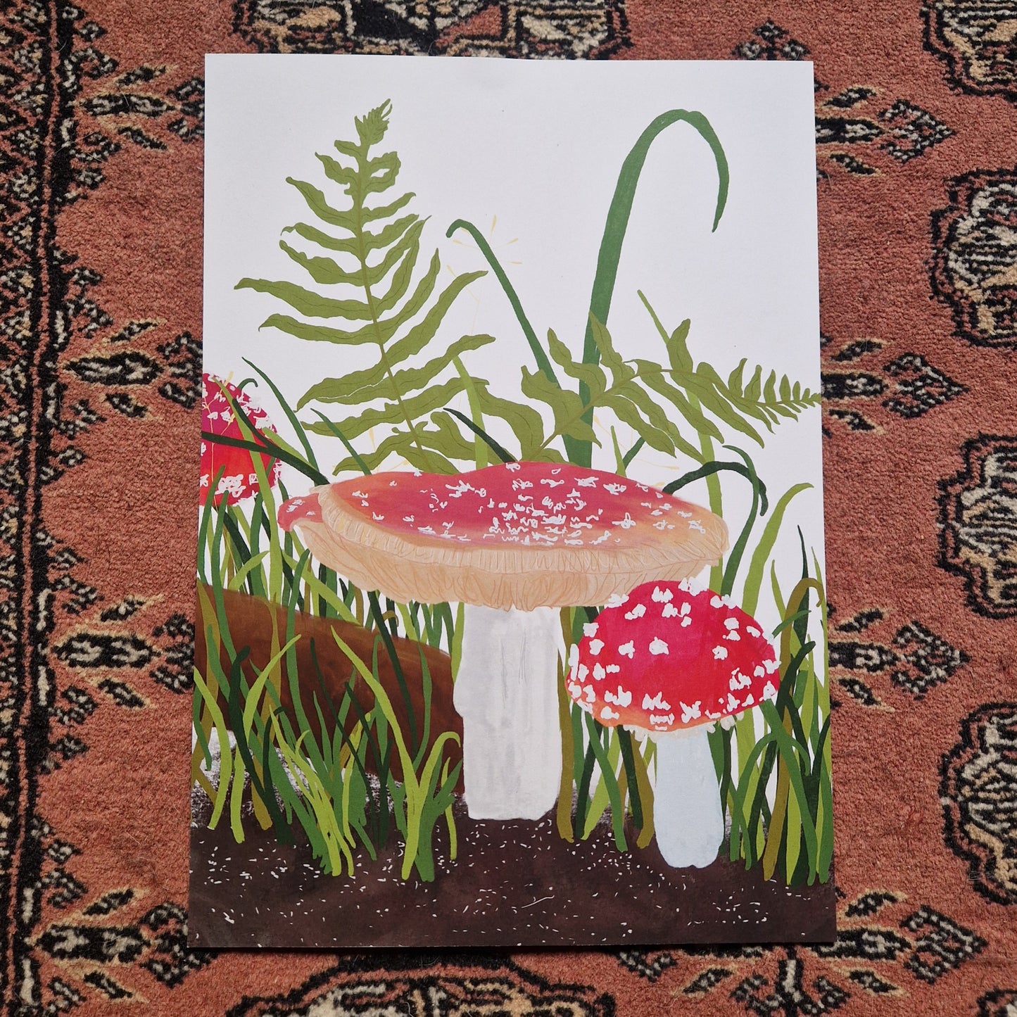 A view of the full print on a textured red patterned background. The print is white with fly agaric fungi growing from the bottom of the print along with ferns and green grasses. Across the bottom of the print you can see a brown soil.