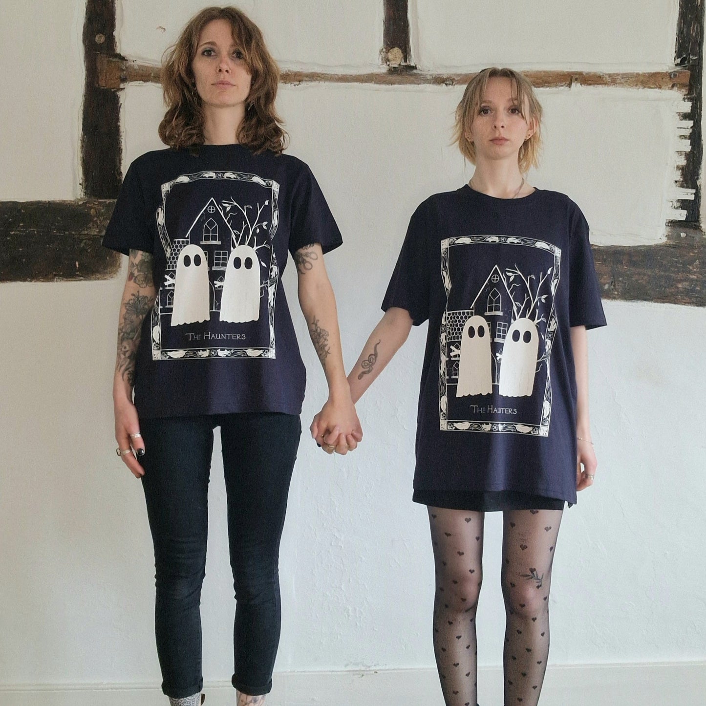 Two people wearing dark navy t-shirts with a graphic design of ghosts in a room with exposed brick walls.