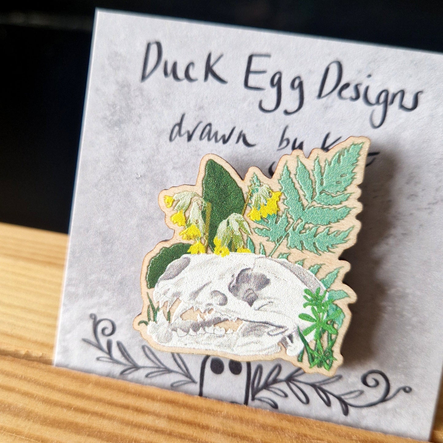 The Badger Skull and Wildflowers Pin Badge from Duck Egg Designs features a wooden badger skull with green leaves and yellow wildflowers, displayed on a branded card and resting on a wooden surface.