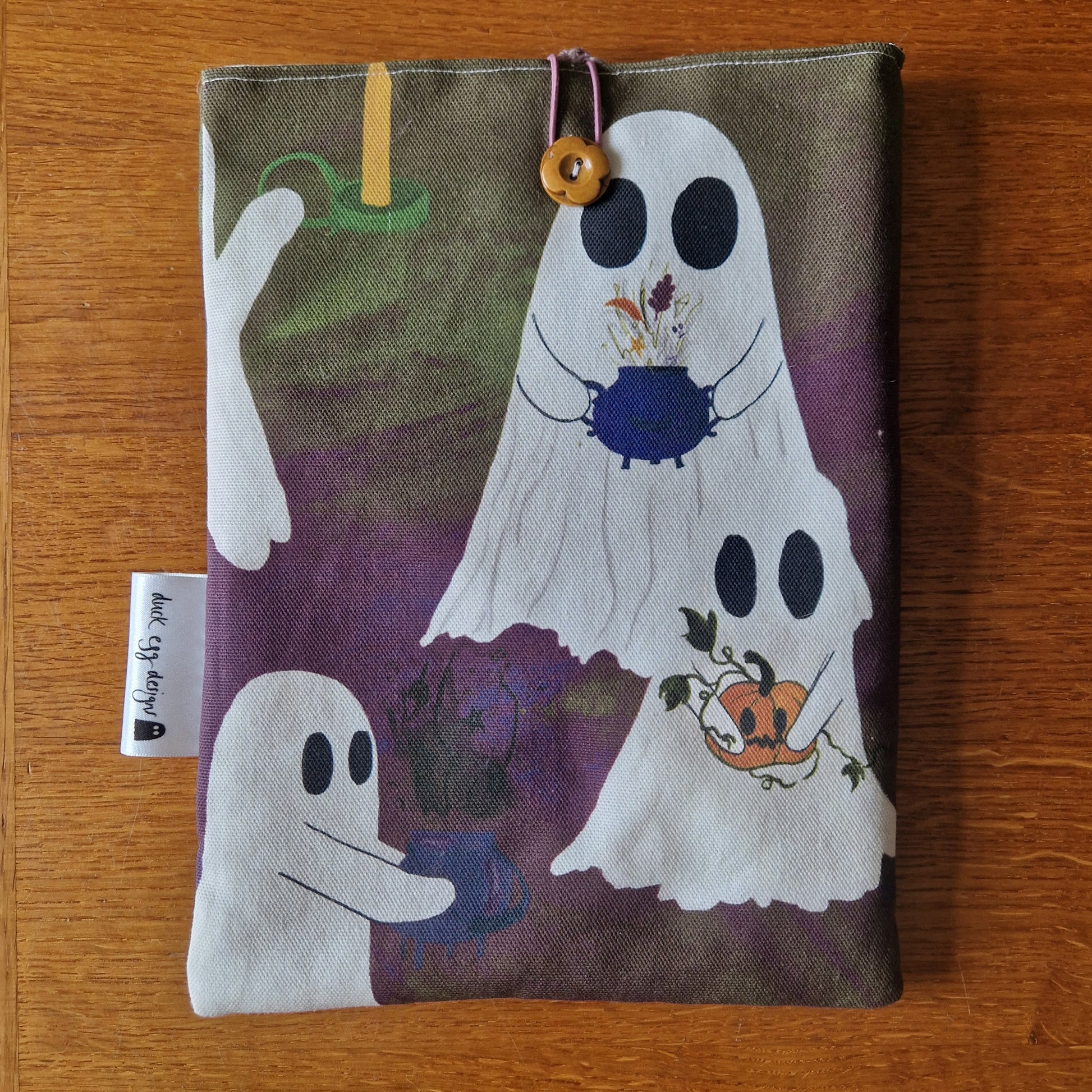 The A Fright Of Ghosts Book Sleeve by Duck Egg Designs features cartoon ghosts performing magic with cauldrons, flowers, and pumpkins on a purple and green background—perfect protection for your favorite books.
