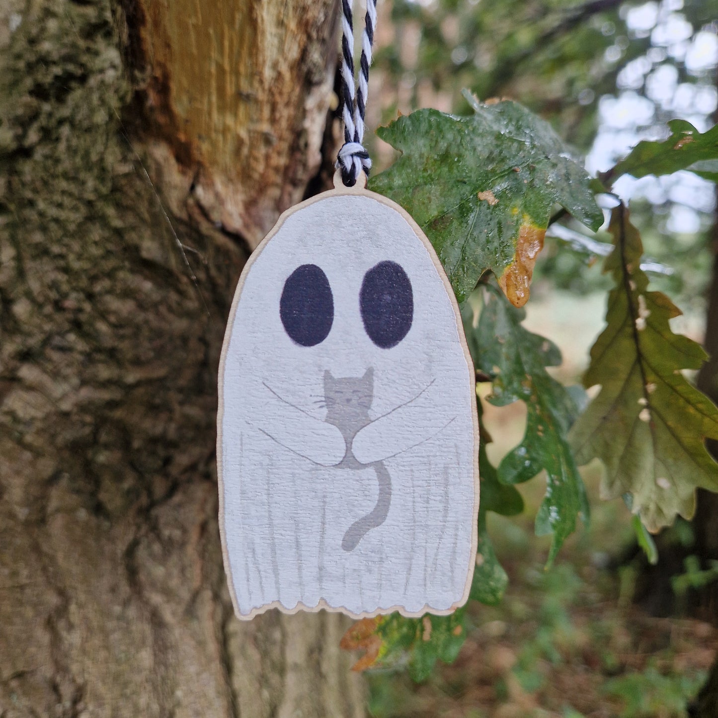 The Ghost With Cat Hanging Wooden Decoration by Duck Egg Designs features a wooden ghost holding a gray cat, hanging from a black-and-white string on a tree branch next to green leaves—perfect for charming wall decor.
