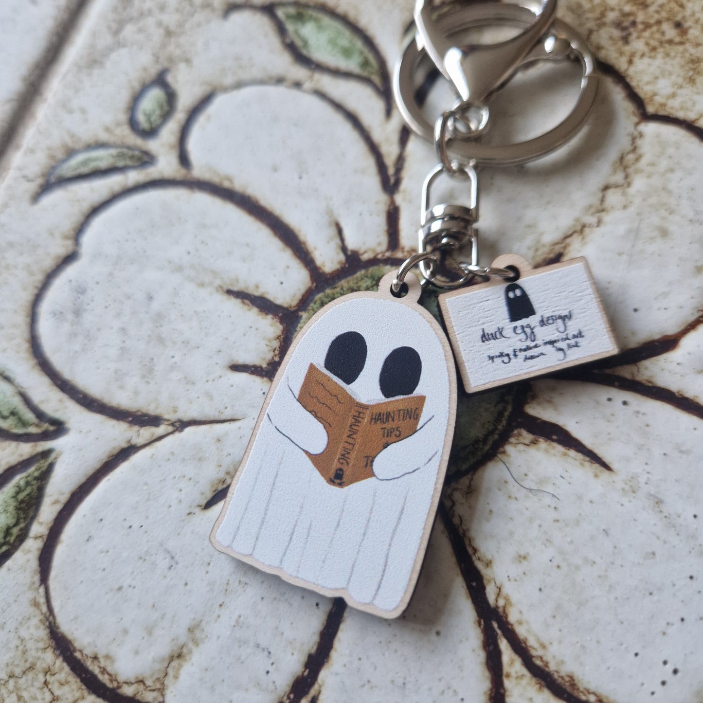 The Reading Ghost Keyring by Duck Egg Designs features a cute cartoon ghost reading "Haunting Tips," with a small notebook charm, shown on a floral-patterned ceramic surface.