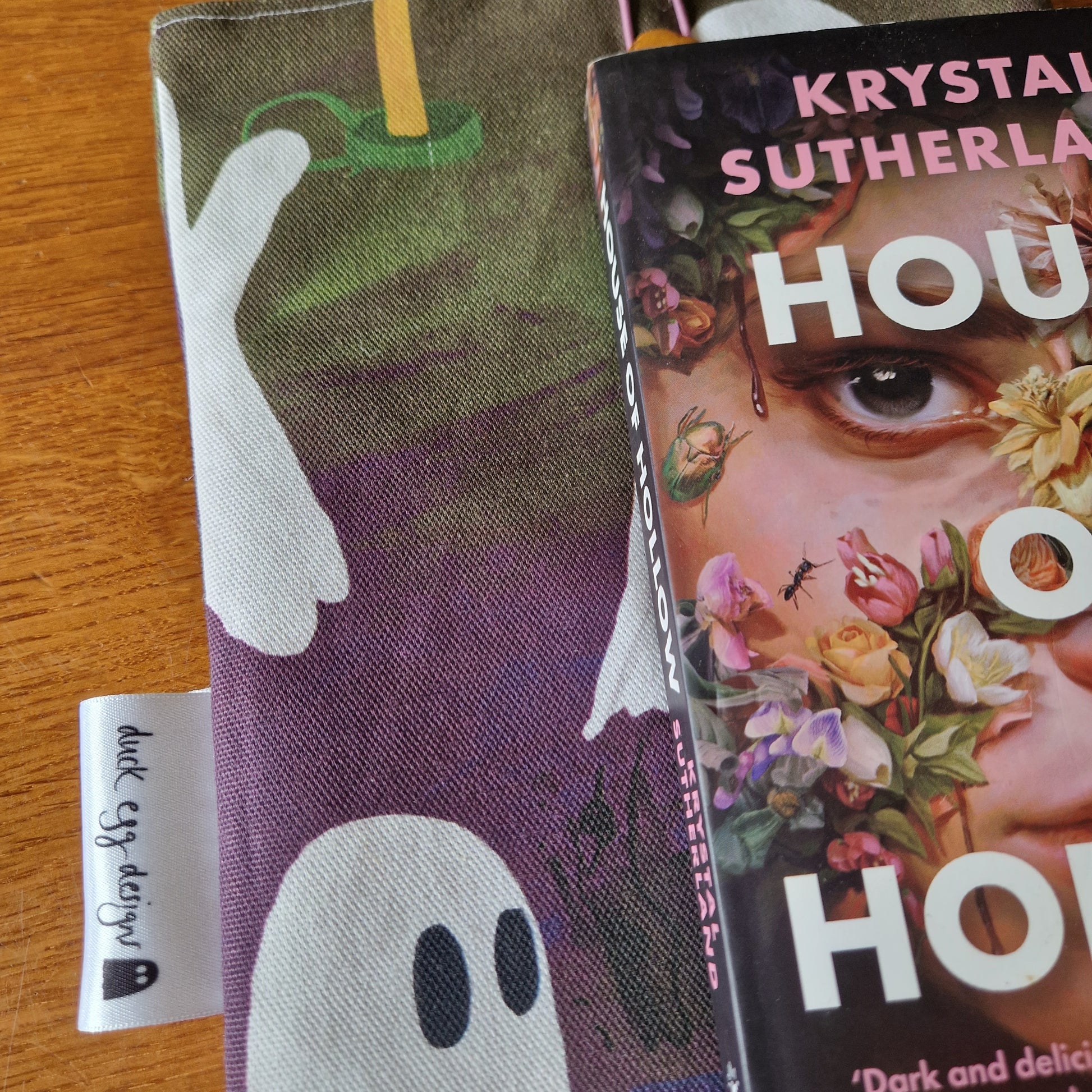 The book House of Hollow by Krystal Sutherland rests partly on the A Fright Of Ghosts Book Sleeve by Duck Egg Designs, featuring ghost illustrations, atop a wooden surface.