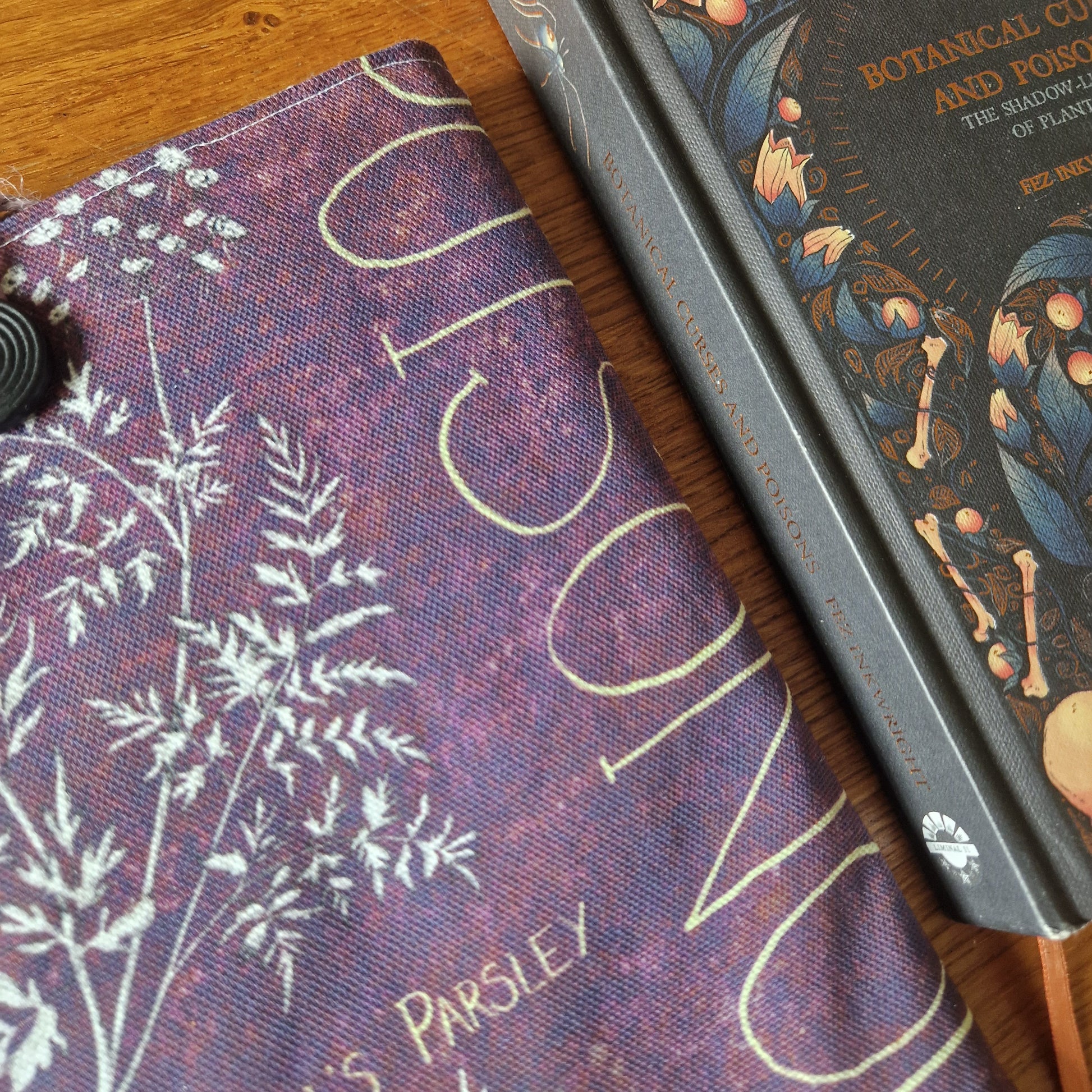 Close-up of two books on wood: one with botanical art, the other with herbs and bold text, partially covered by Duck Egg Designs handmade Poison Plants Book Sleeve—crafted from upcycled fabric with a black elastic cord.