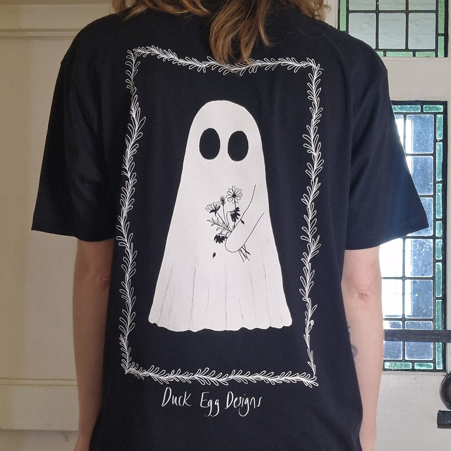 Person wearing a black t-shirt with a white ghost holding flowers design on the back, standing indoors.