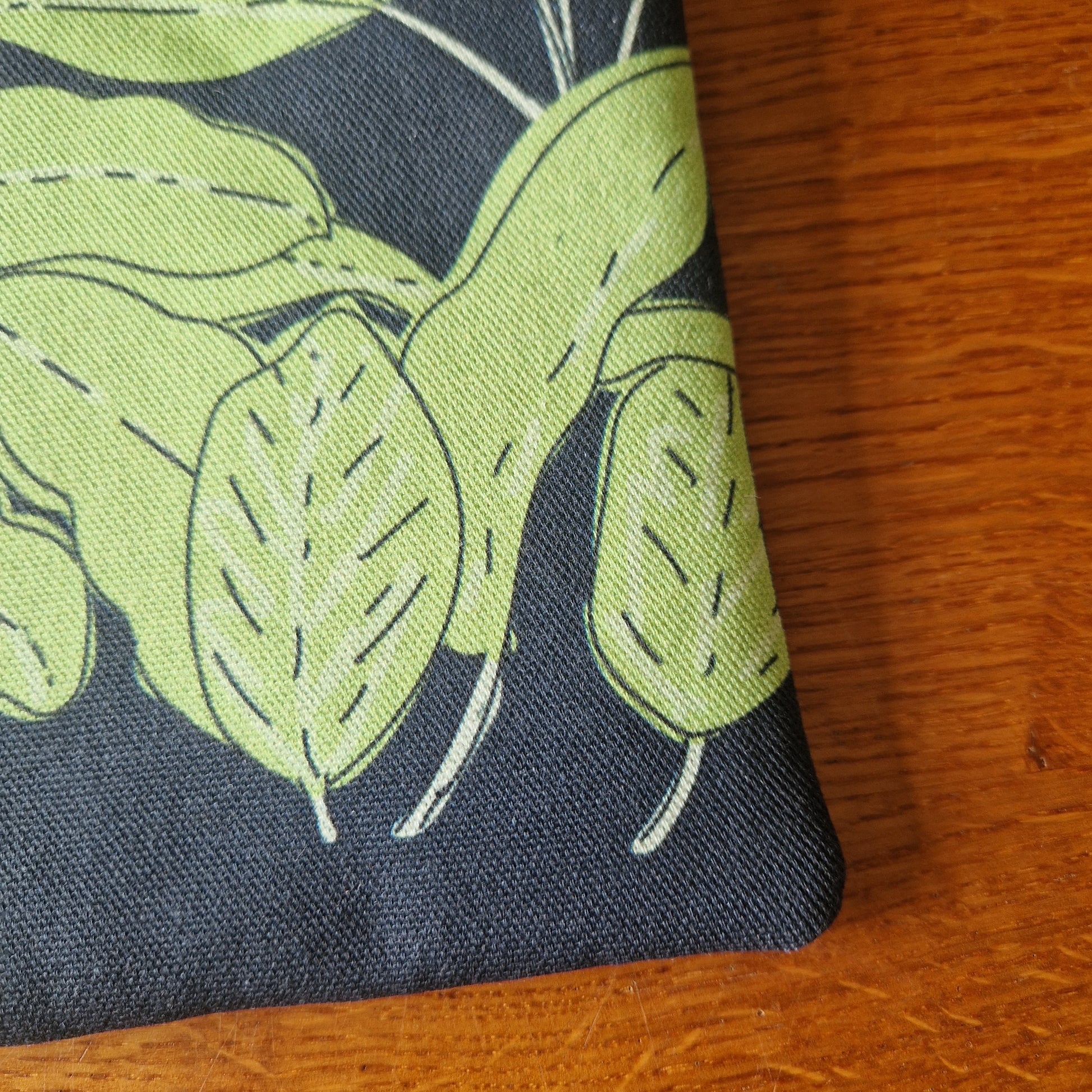 A close-up of the Common Herbs Kindle Pouch by Duck Egg Designs, handmade with a green plant pattern on dark fabric and stitched leaf and bean pod outlines, displayed on a wooden surface.