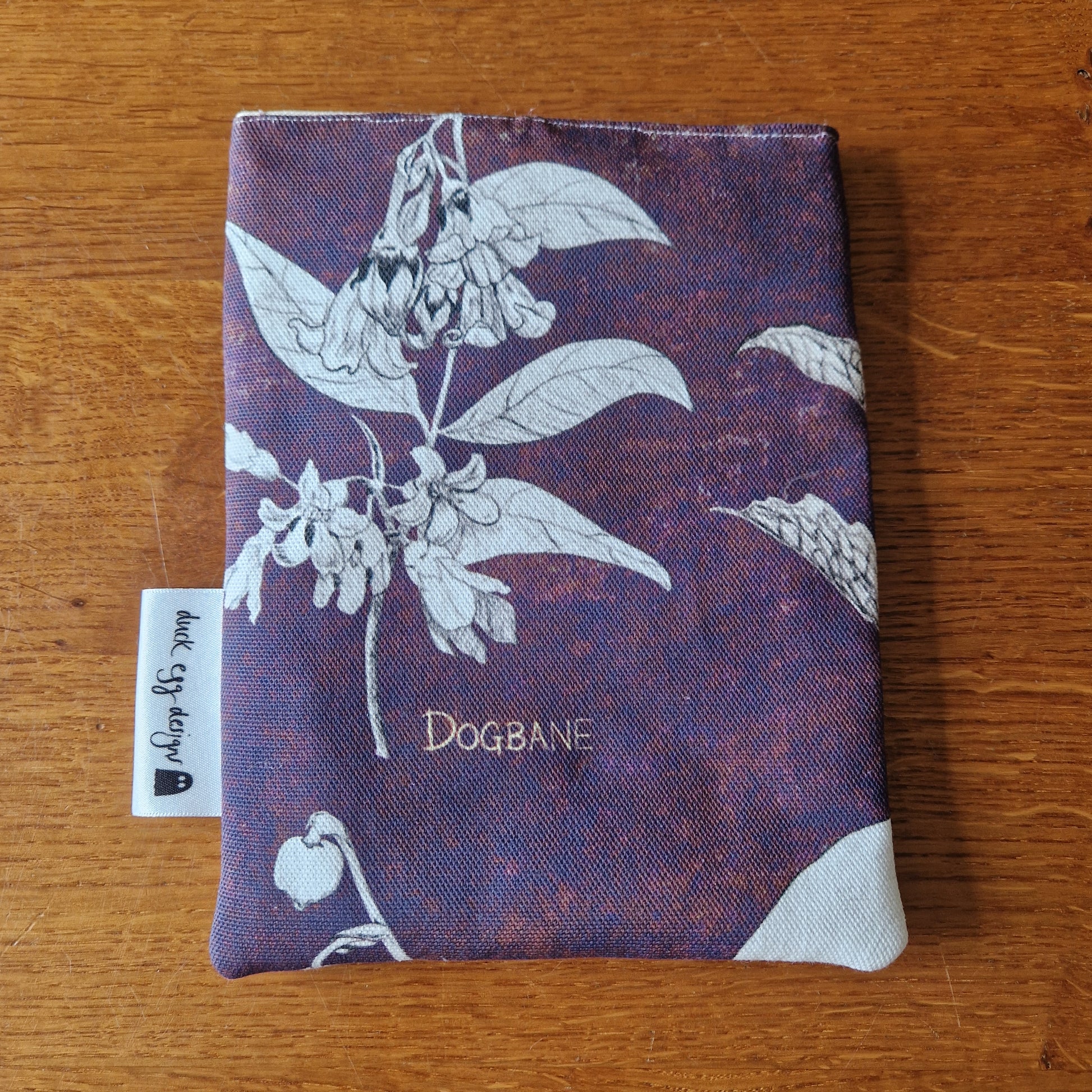 The Poison Plants Kindle Pouch by Duck Egg Designs features a purple fabric with white botanical illustrations and DOGBANE text, plus a small side tag—an ideal unique Kindle sleeve for book lovers.