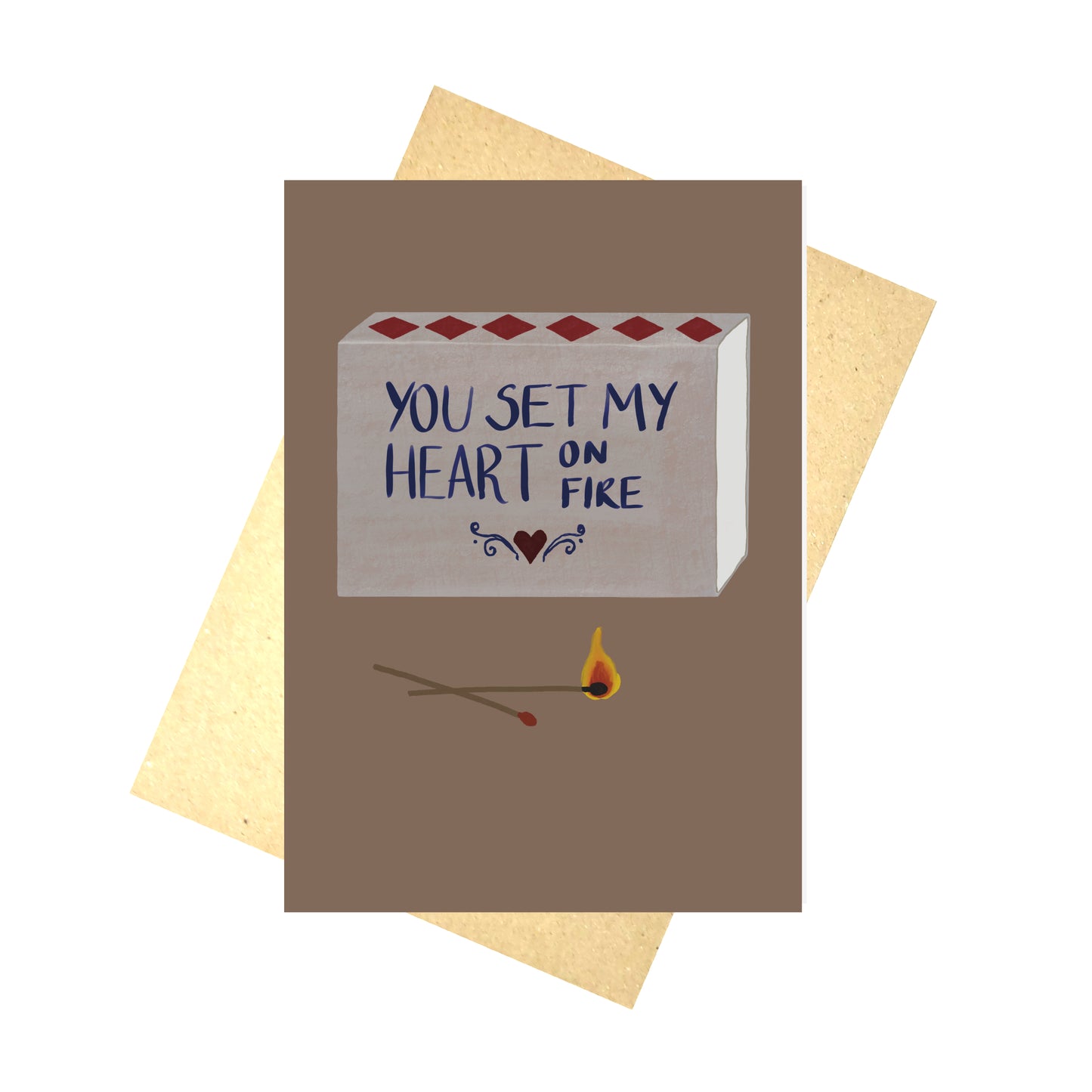 A warm browny purple card with a grey matchbox design on it sits in front of a brown recycled paper envelope on a white background. The match box has words on it that read ‘ You set my heart on fire’ in blue with a red heart and blue symmetrical swirls below it. In front of the matchbox are two matches, one of which is on fire.