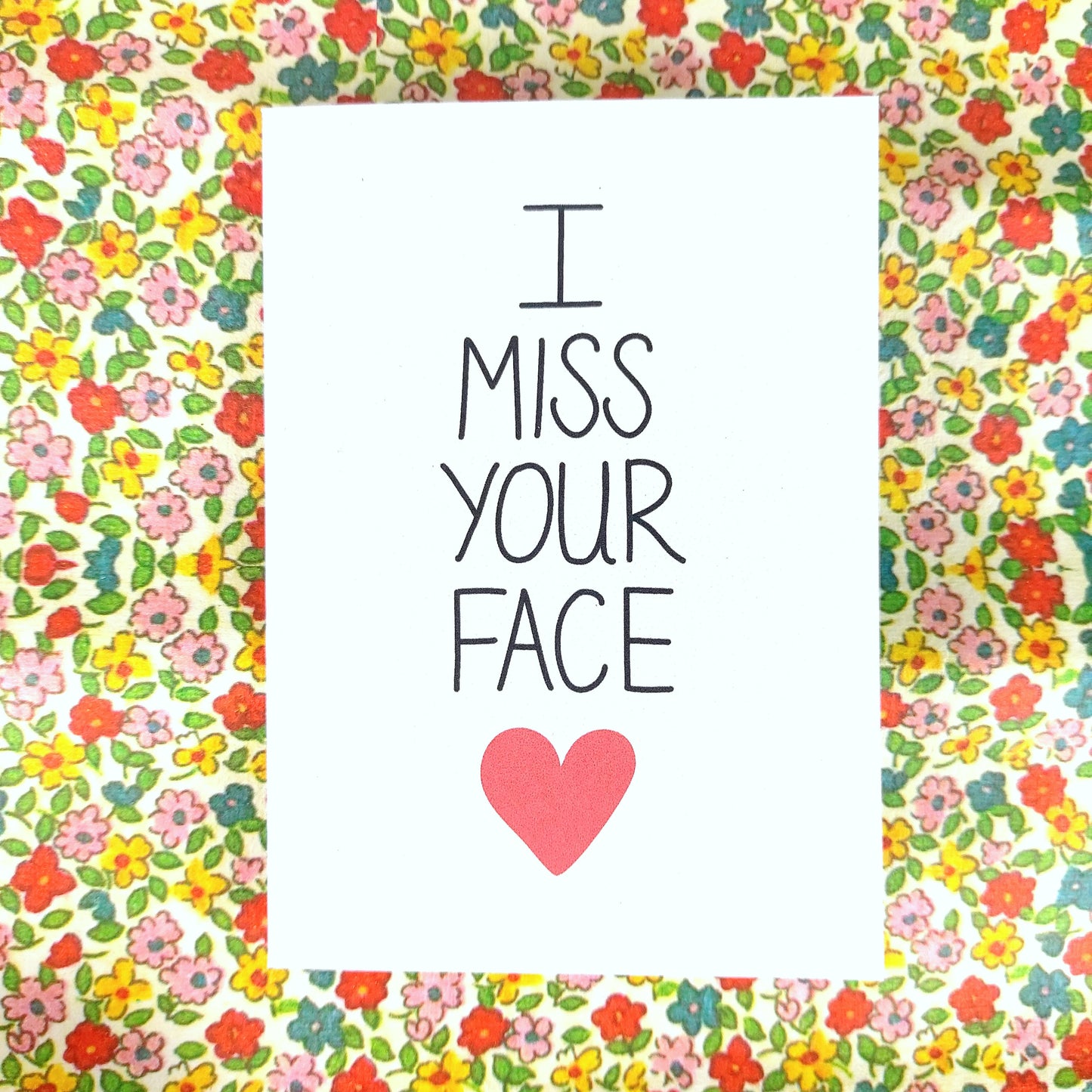 A white card with the words 'I MISS YOUR FACE' down the middle, below which is a red heart. Behind the card is a floral patterned background.