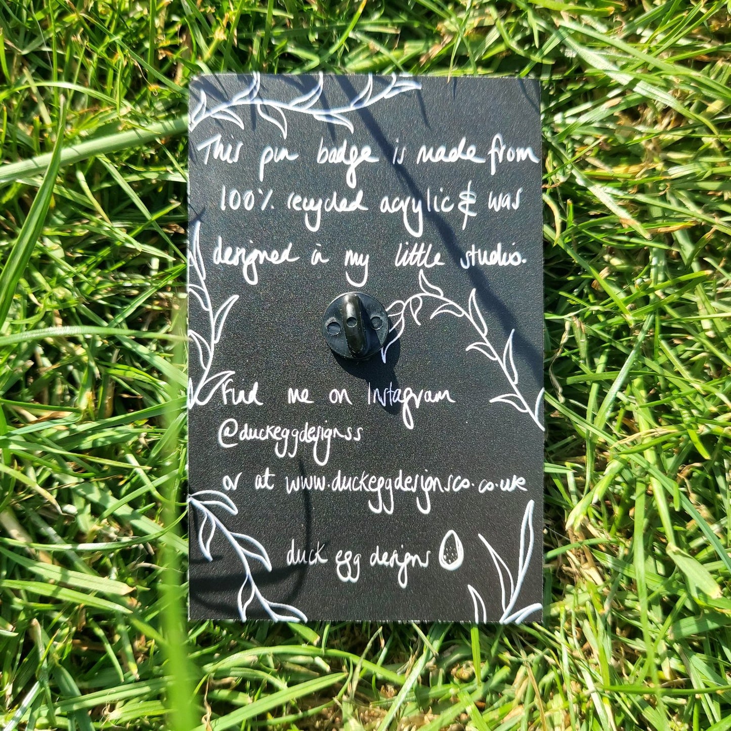 The back of the black backing card featuring information about the pin badges in white handwriting, as well as the outline of leafy vines. Behind the backing card is a green grassy background.