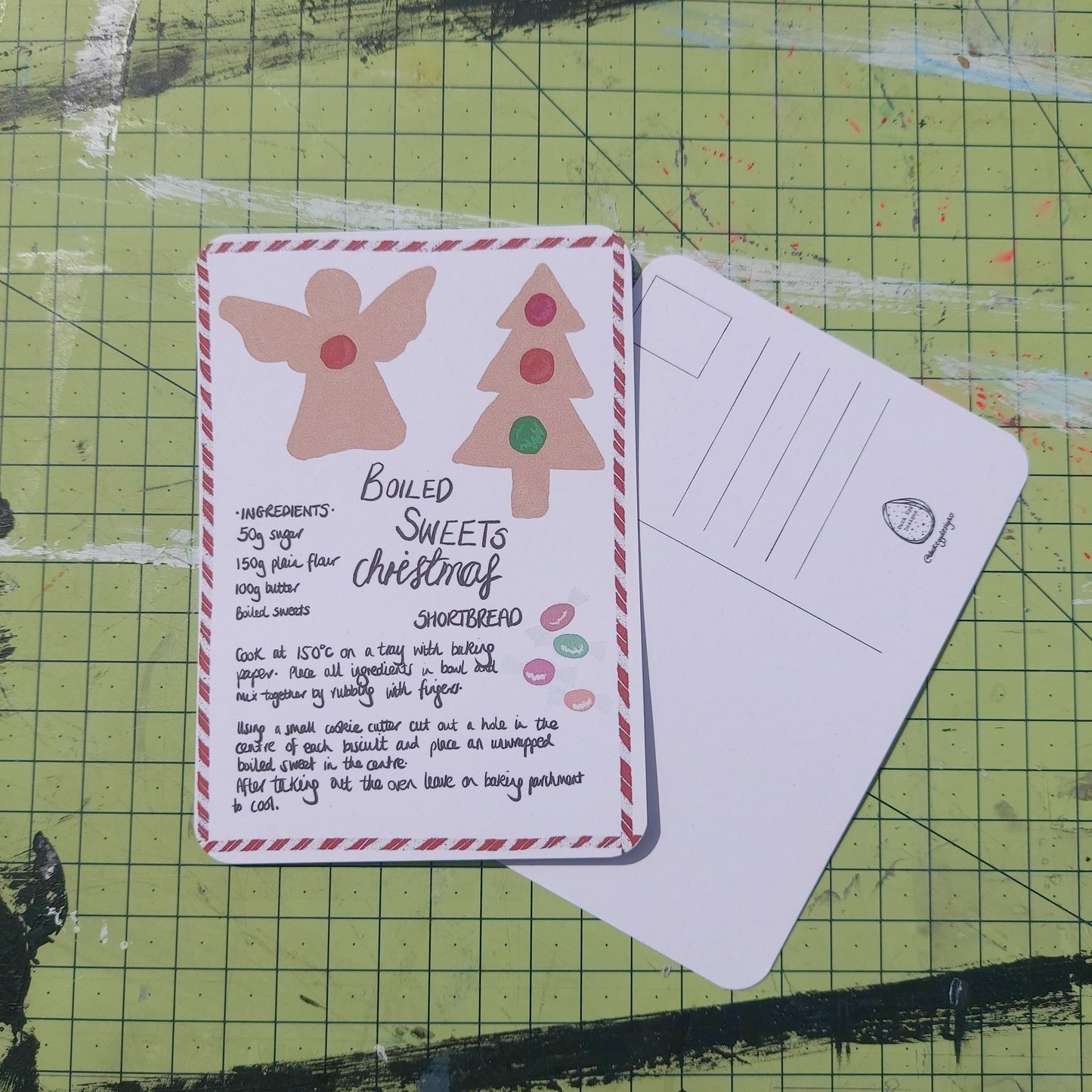 Front and back of a postcard shown on a green cutting mat. The front shows a handwritten boiled sweets Christmas shortbread recipe bordered by red and white candy cane, with angel and Christmas tree biscuits on the card.