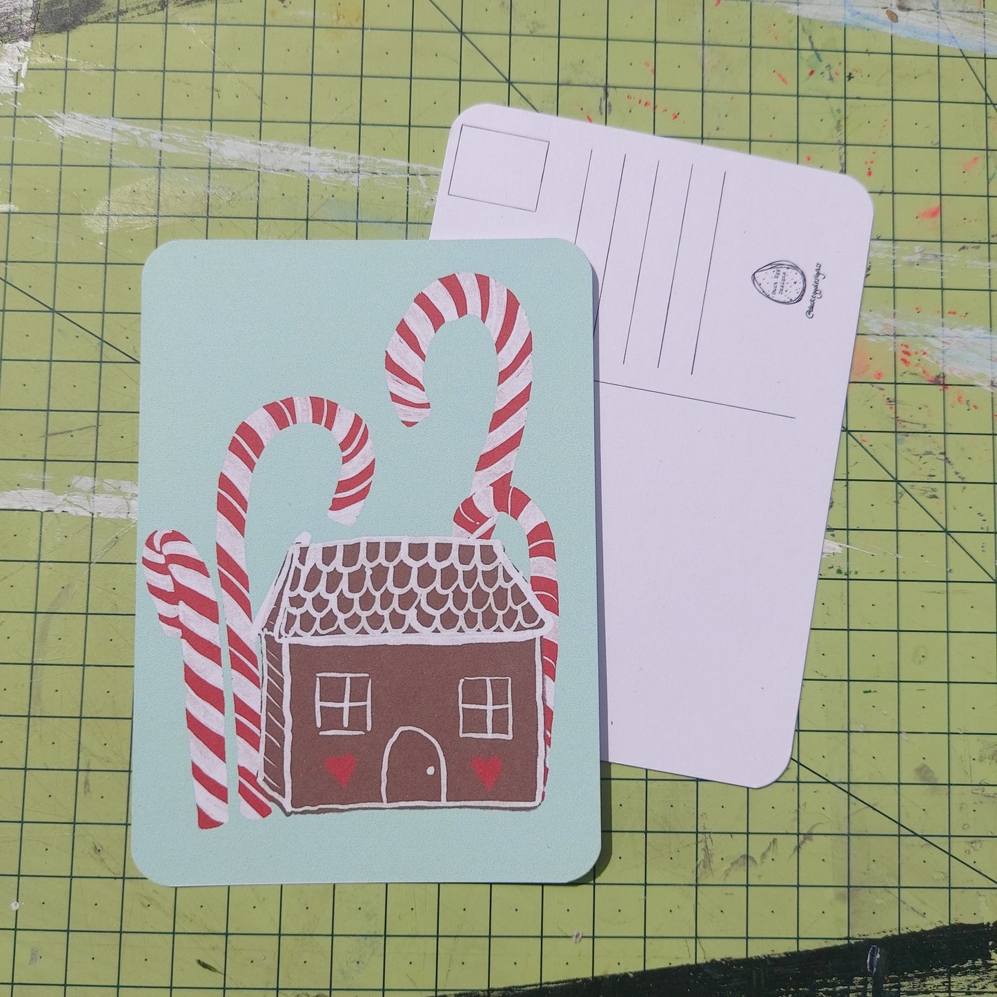 Front and back of a postcard are visible on a green cutting mat. Front of the postcard shows a gingerbread house with red and white icing under some tall red and white candy canes on a peppermint green background. The back of the postcard is white, featuring lines for the address and a square for the stamp.