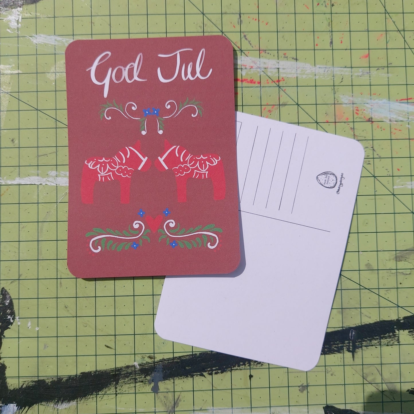 Front and back of a postcard are visible on a green cutting mat. The front of the postcard is red with two red dala horses with the words ‘God Jul’ above them in red and abstract curving shapes underneath them. The back of the postcard features lines for the address, a square for the stamp and the duck egg design logo.