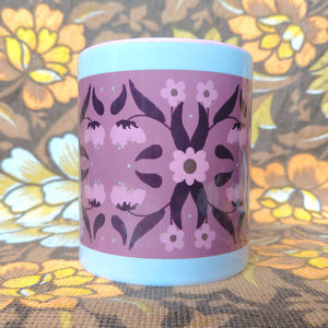 Pink and white mug with a symmetrical floral retro inspired pattern sits in front of a white and brown floral background. The mug features a pink handle and inside, with a border of white around the design.