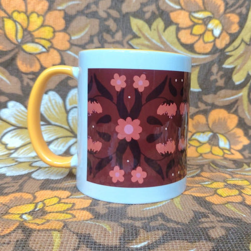 A white mug featuring a yellow handle and inside sits on a brown and white floral fabric. The mug features a brown rectangle with a symmetrical retro inspired floral and leaf pattern in orange and dark brown.