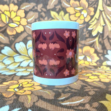 Load image into Gallery viewer, A white mug featuring a yellow handle and inside sits on a brown and white floral fabric. The mug features a brown rectangle with a symmetrical retro inspired floral and leaf pattern in orange and dark brown
