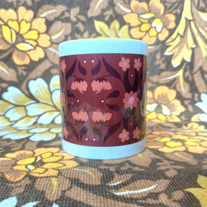 A white mug featuring a yellow handle and inside sits on a brown and white floral fabric. The mug features a brown rectangle with a symmetrical retro inspired floral and leaf pattern in orange and dark brown