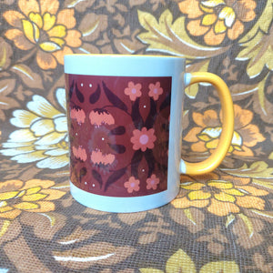 A white mug featuring a yellow handle and inside sits on a brown and white floral fabric. The mug features a brown rectangle with a symmetrical retro inspired floral and leaf pattern in orange and dark brown