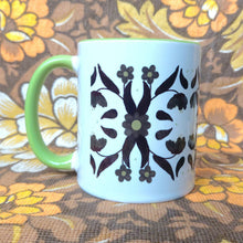 Load image into Gallery viewer, A white mug with a green handle and inside sits in front of a brown and white floral background. The mug features a symmetrical 70s inspired floral pattern in brown and green.
